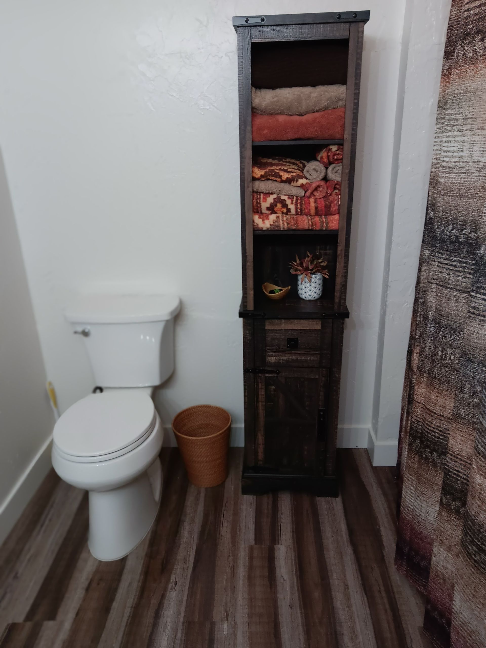 A white toilet sits next to a tall, dark wood storage unit filled with folded towels and toiletries in a bathroom.