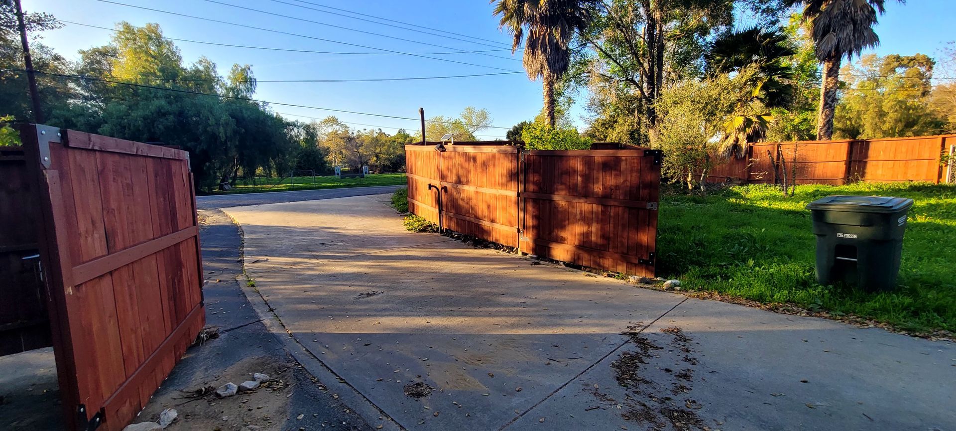 A paved driveway leads toward a wooden fence and trees under a clear, sunny sky, with a green bin on the right.