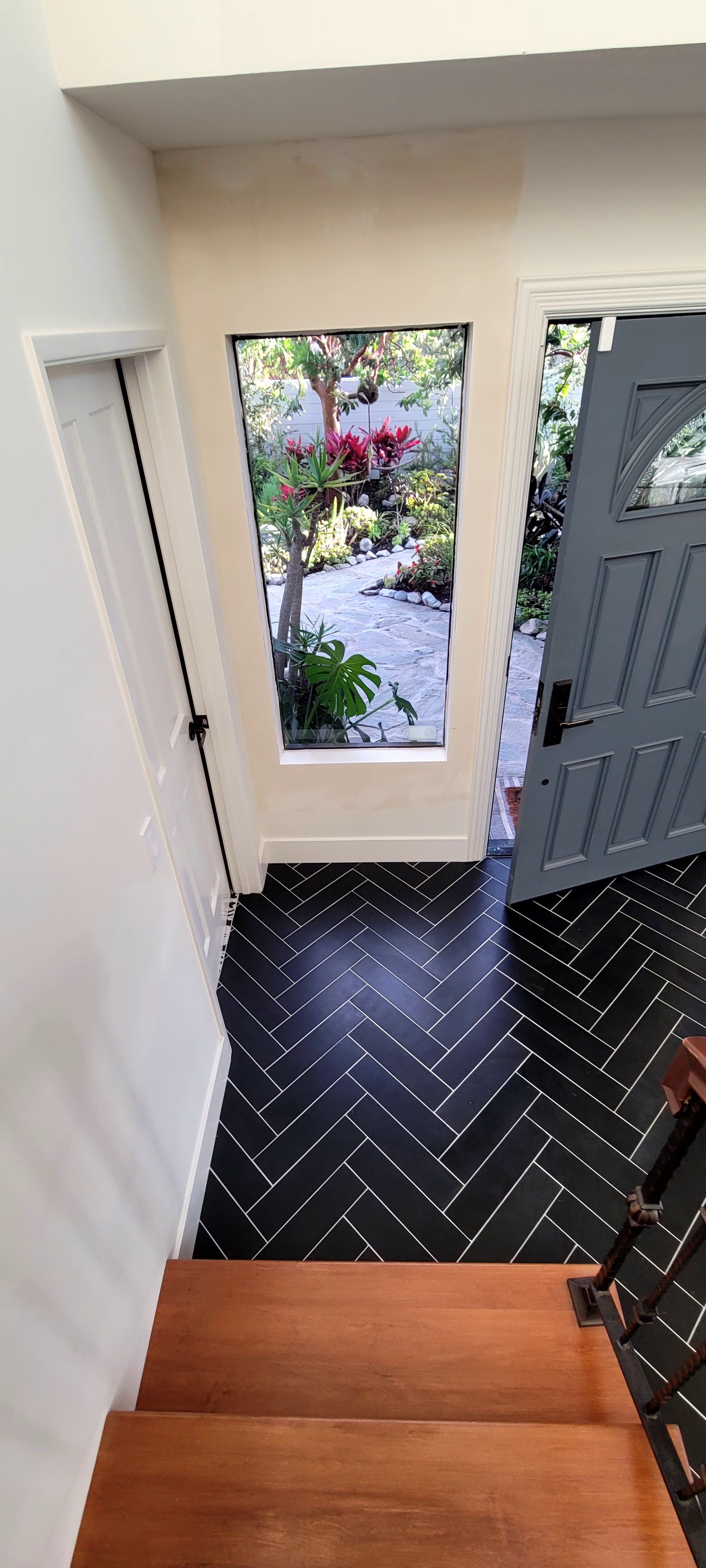 View from wooden stairs looking down at a gray front door and a windowed entryway with black herringbone tile flooring.