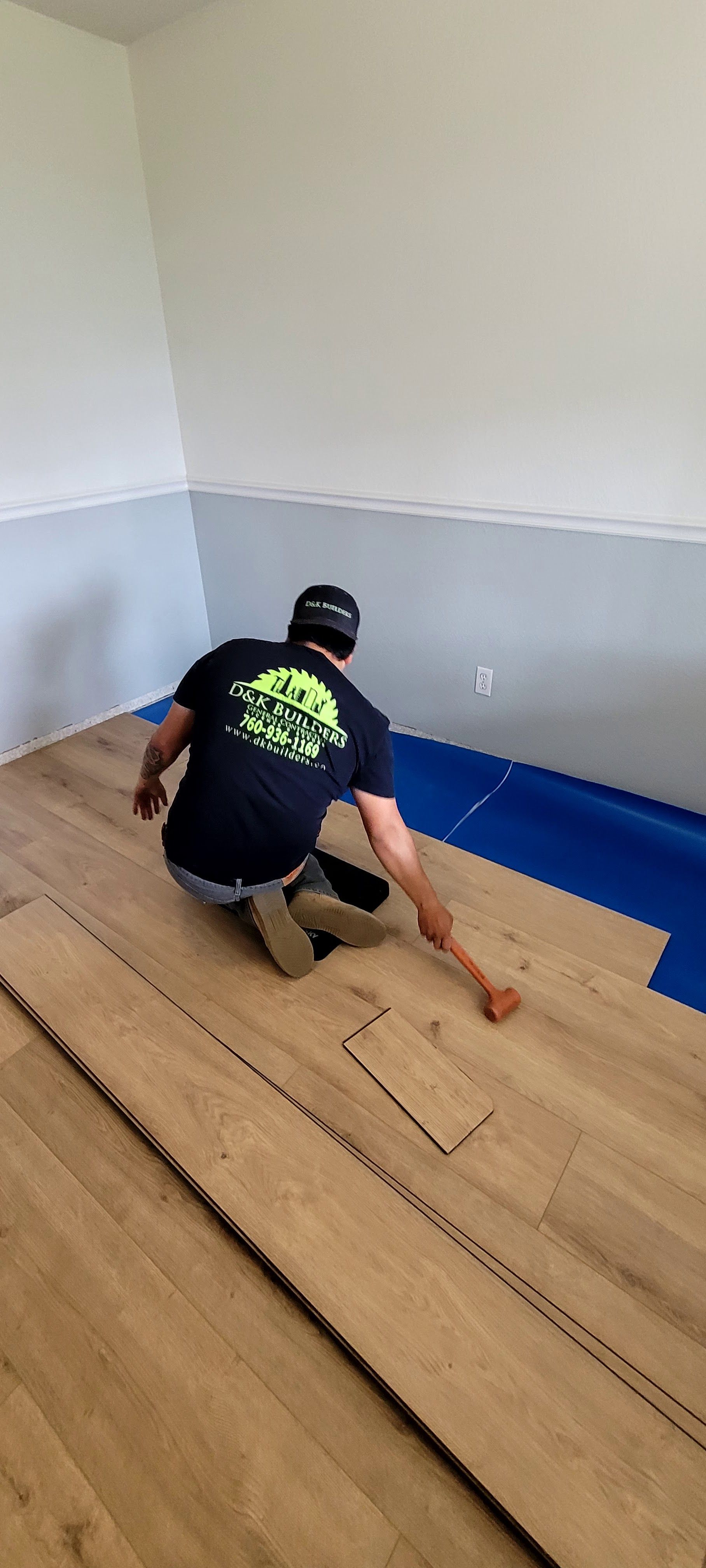A person in a black t-shirt installs light-colored wood laminate flooring over a blue underlayment in a room with grey walls.