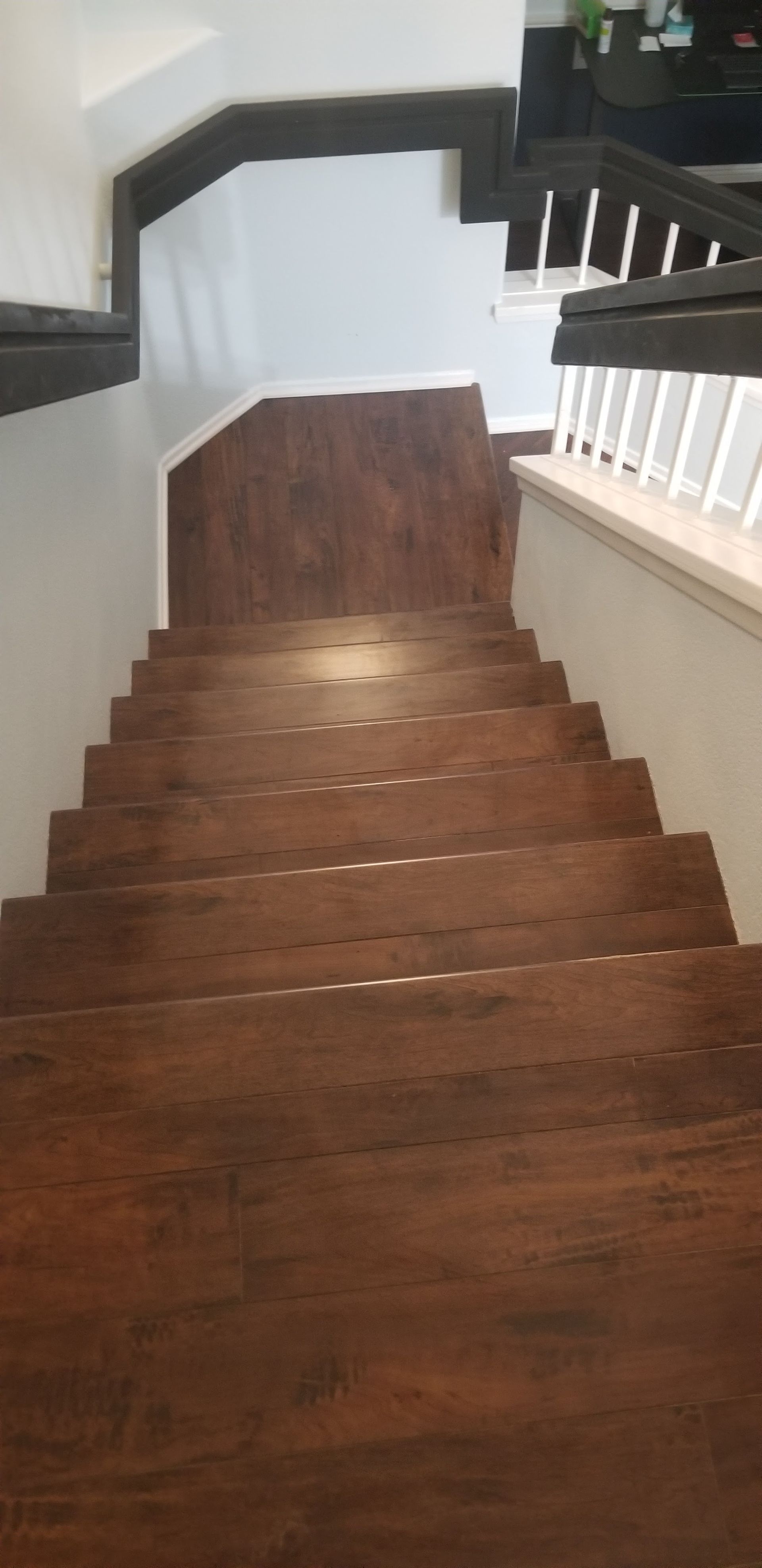 A top-down view of a staircase with dark wood-grain steps leading down to a landing with white walls and a white railing.
