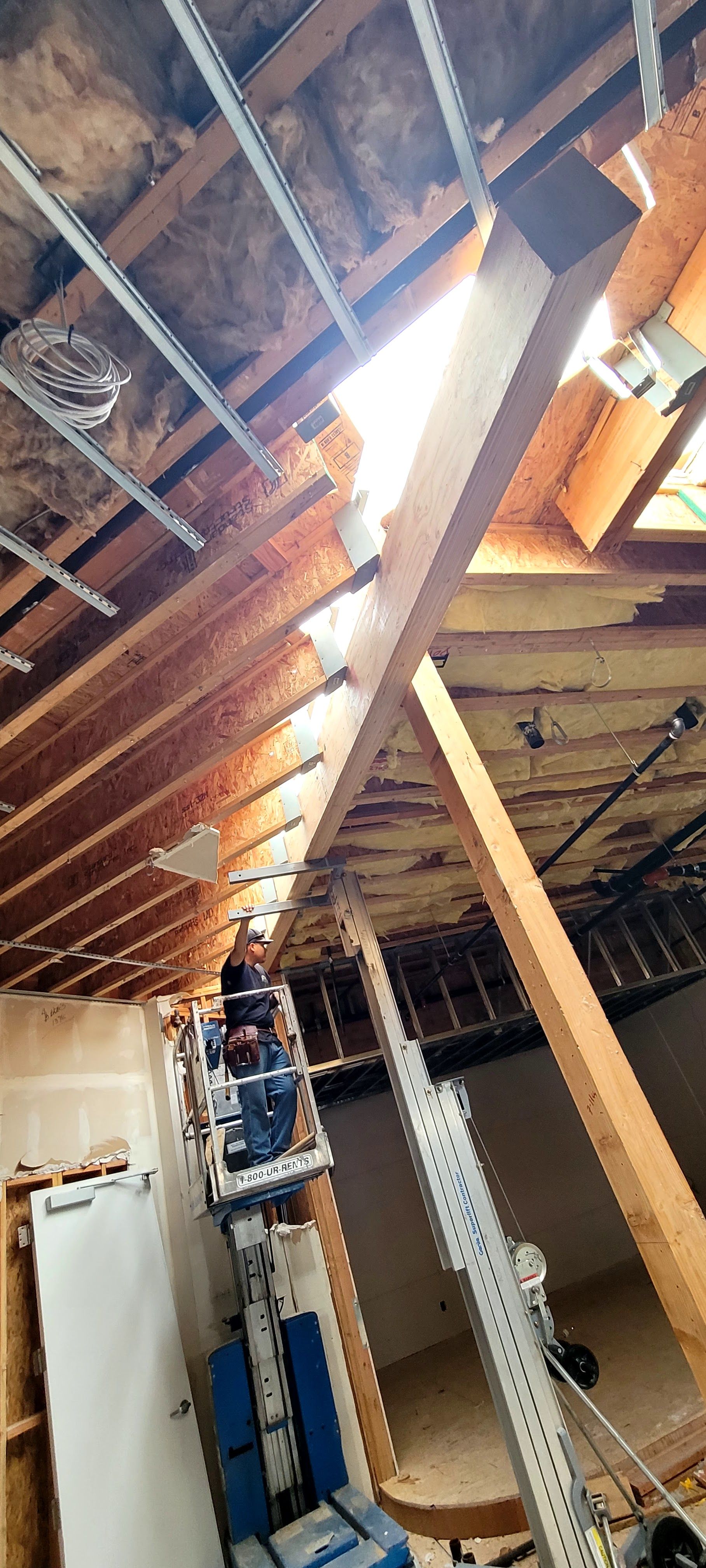A worker on a mechanical lift repairs a damaged wooden ceiling joist system in an unfinished attic space.