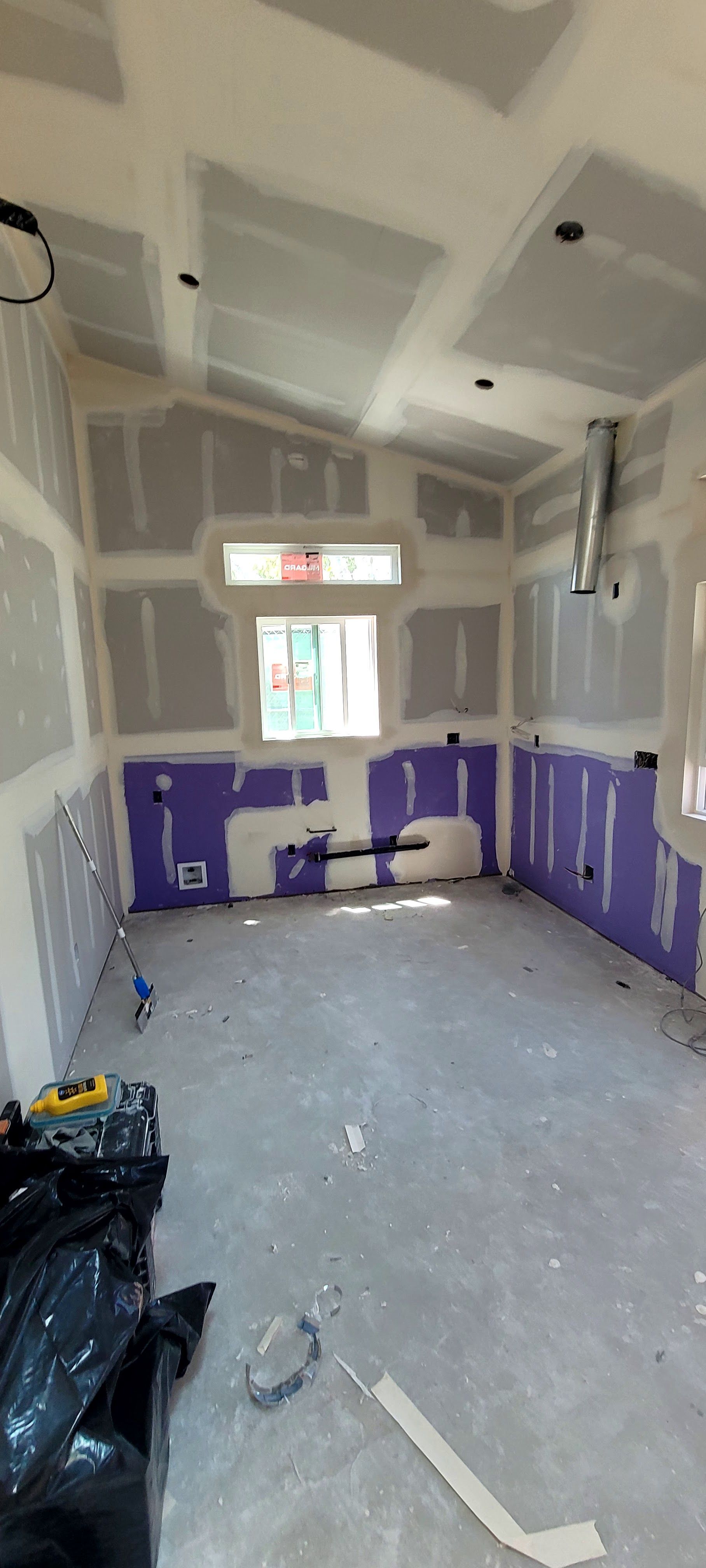 An unfinished room with gray drywall, purple moisture-resistant boards on lower walls, a window, and exposed ductwork.