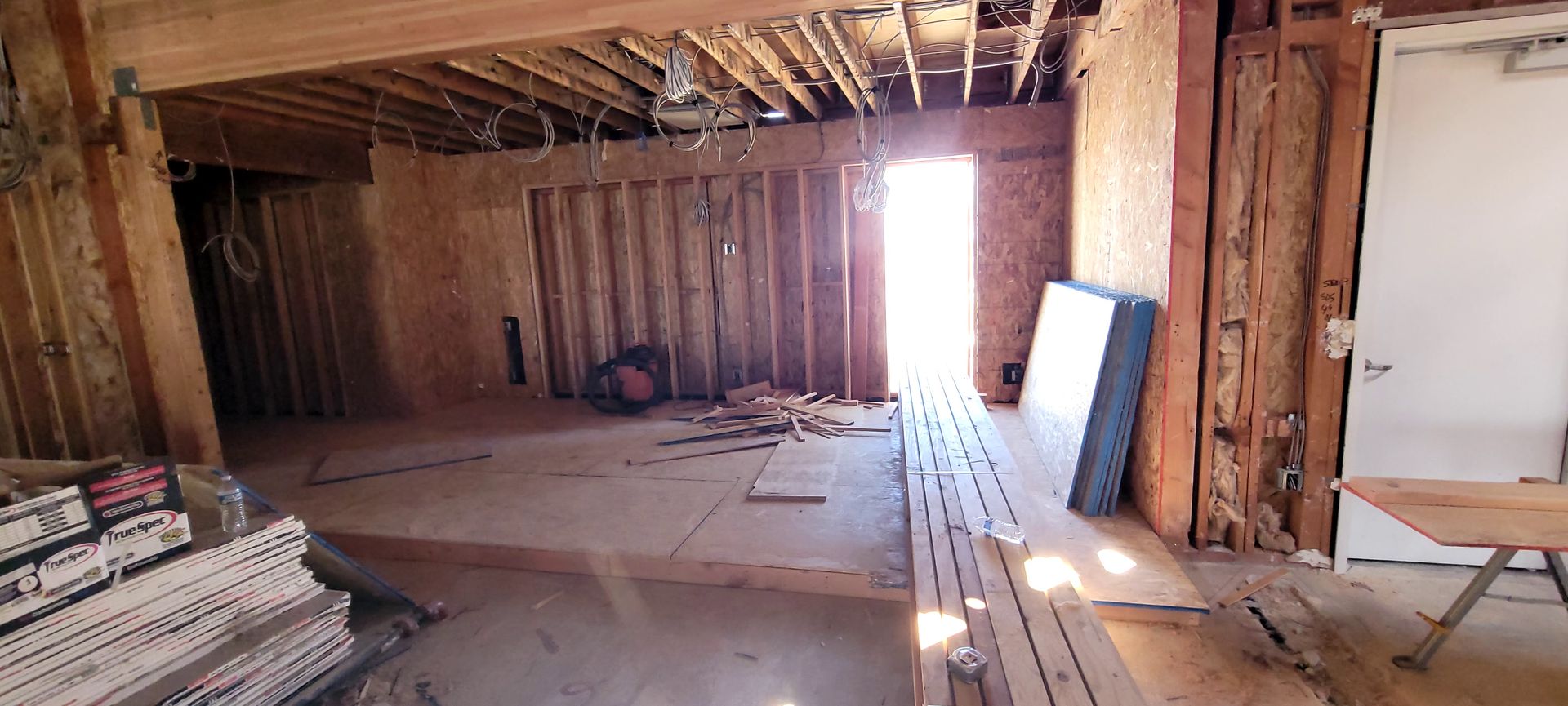A room under construction with exposed wooden wall studs, a plywood floor, unfinished ceiling, and a bright doorway.