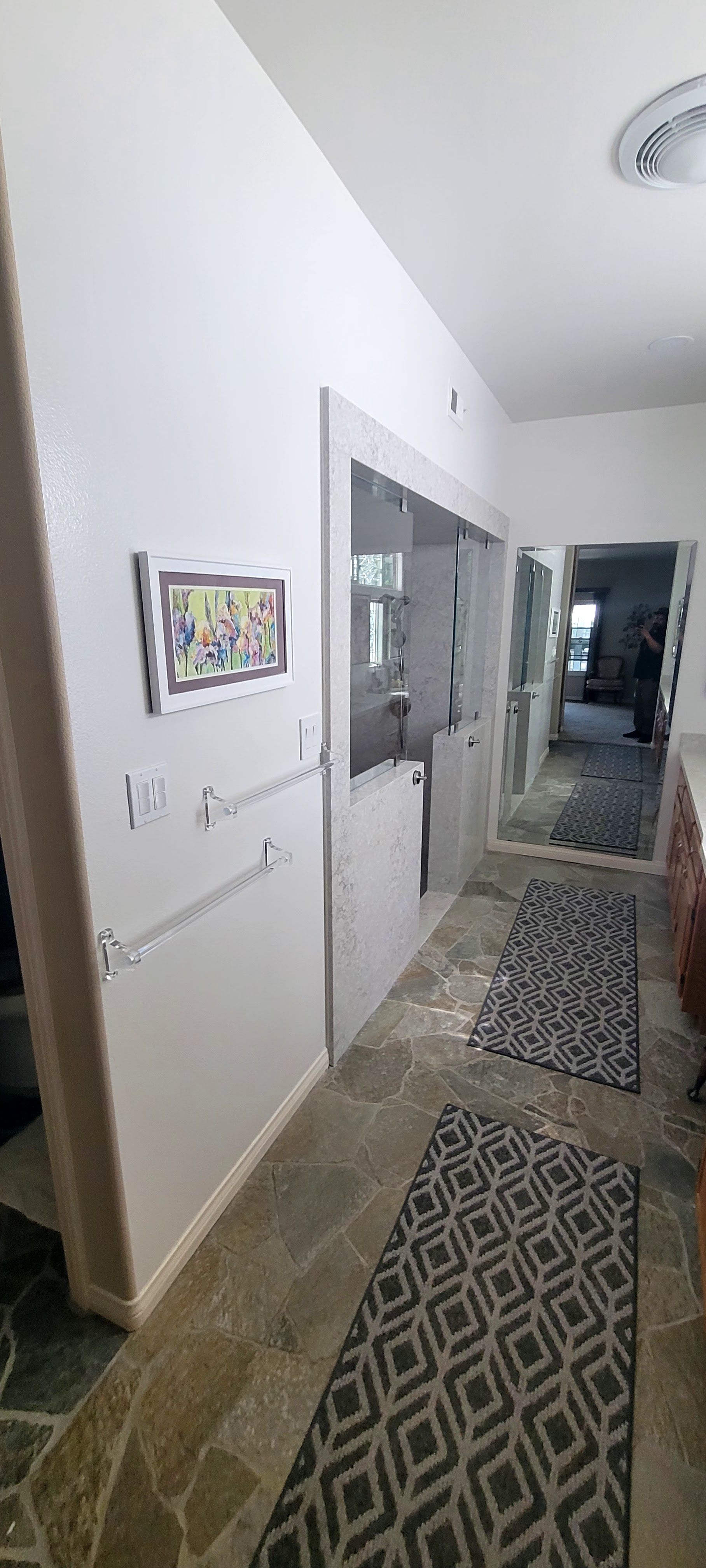 A narrow hallway with stone tile floors, two geometric patterned runners, and a framed art piece hanging on the left wall.