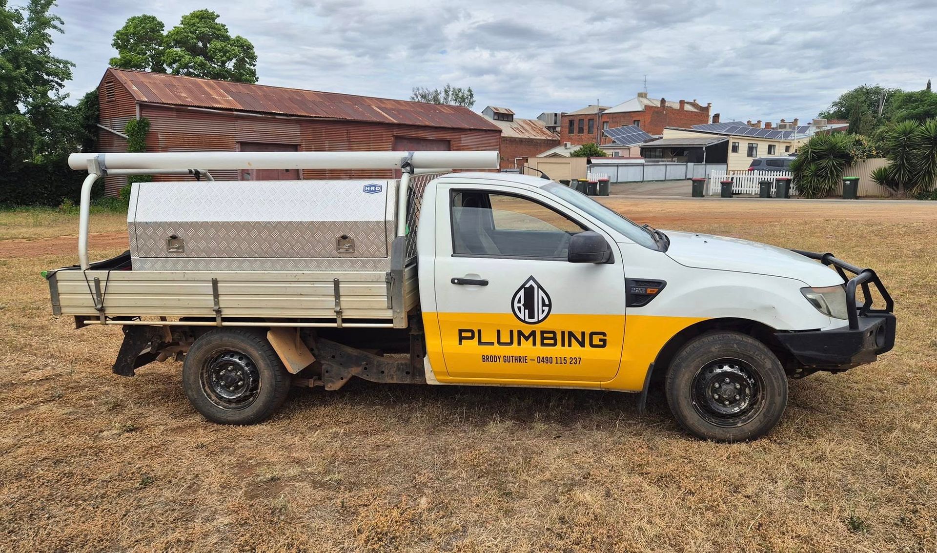 White and yellow plumbing service truck parked on dry grass; a building and trees in background — BJG Plumbing in Coolamon, NSW
