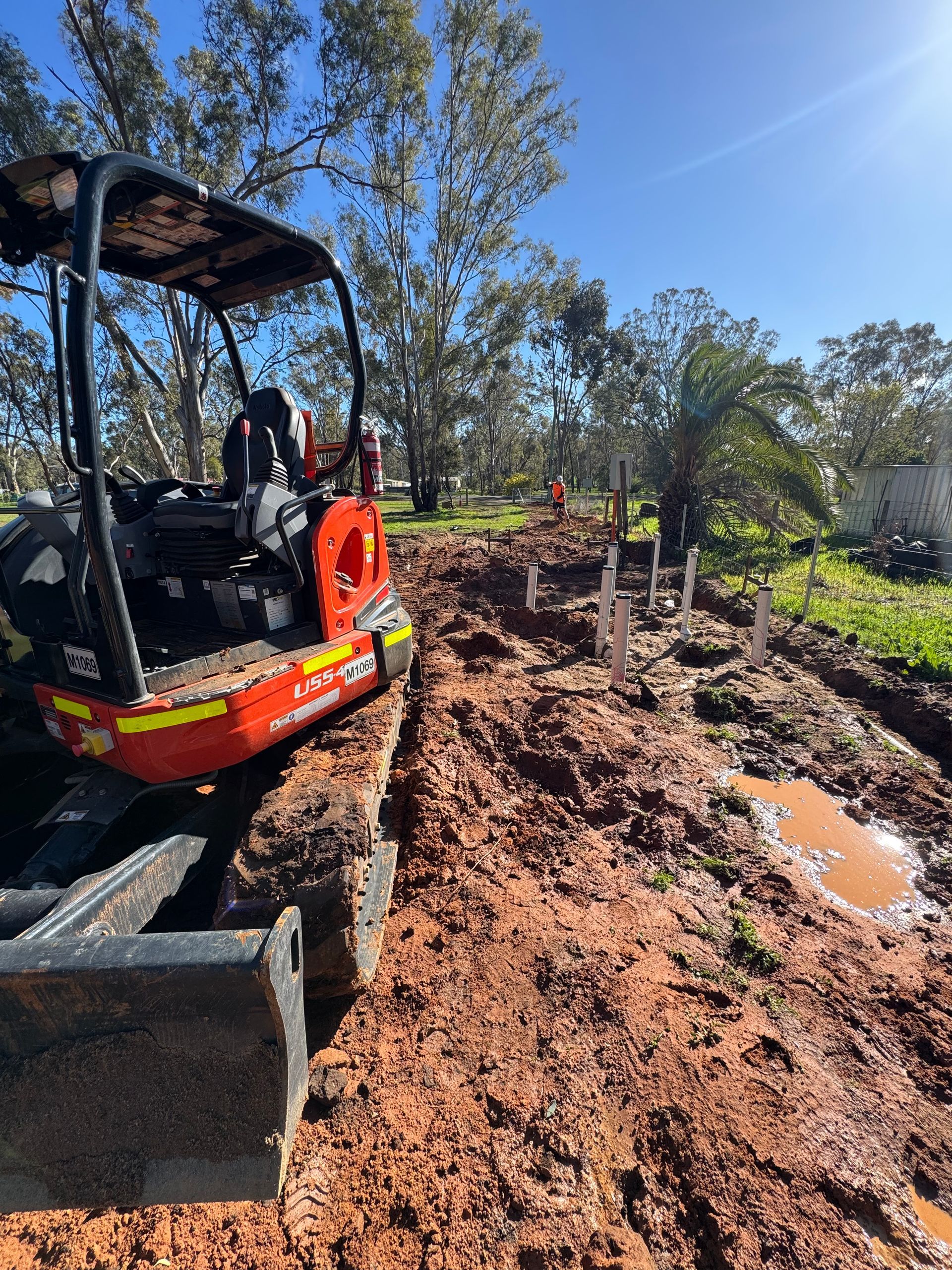 Mini excavator on muddy ground, digging near posts in an outdoor setting with trees under a blue sky - BJG Plumbing
