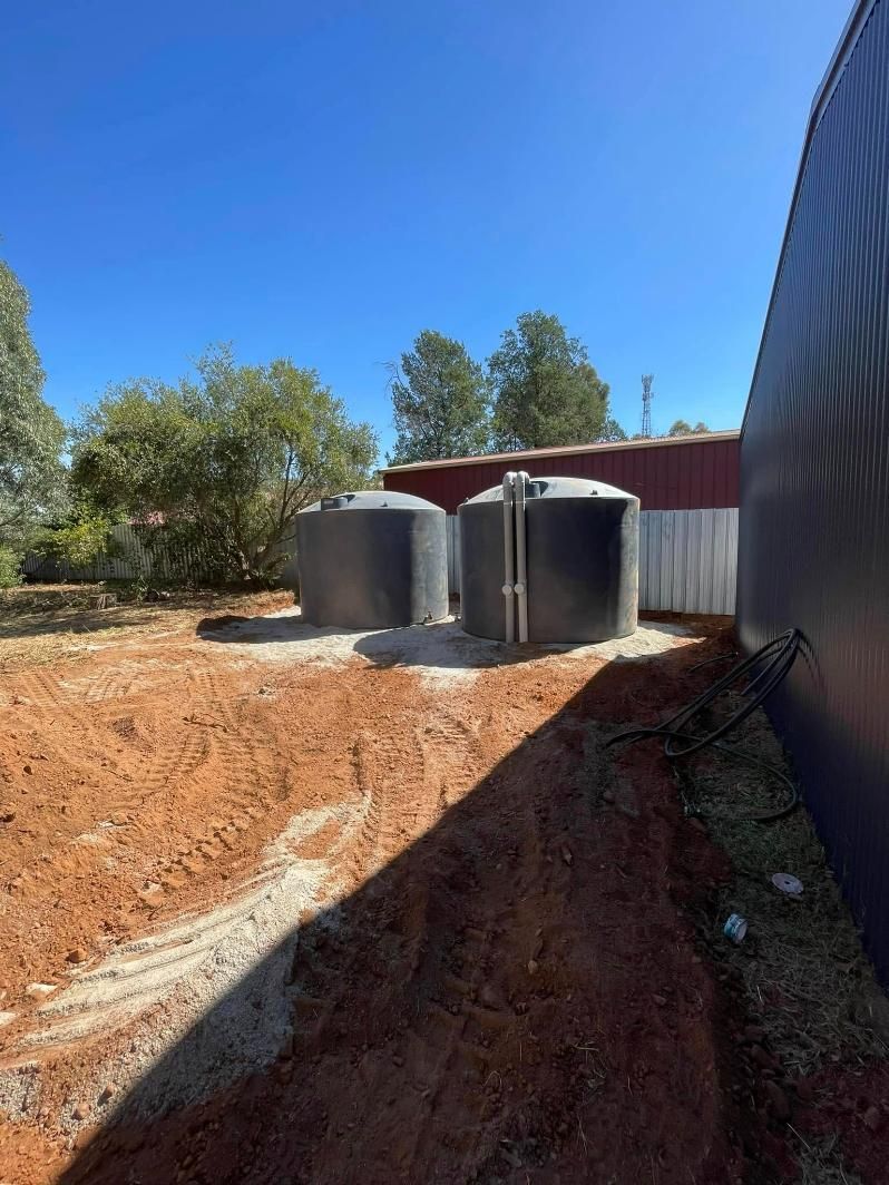 Two dark water tanks next to a building in a dirt yard under a blue sky — BJG Plumbing in Coolamon, NSW