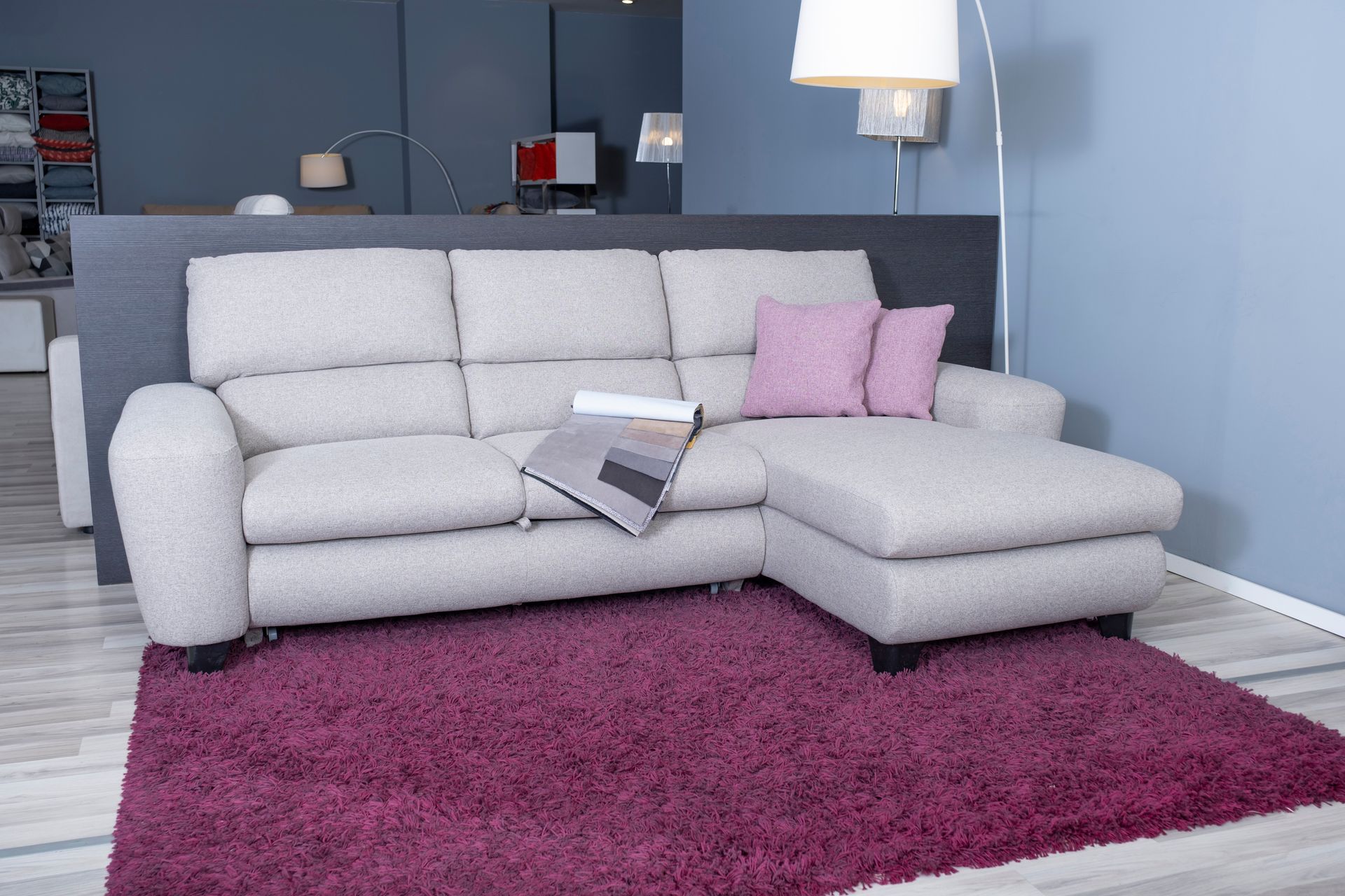 Divano componibile beige con chaise longue su tappeto viola, due cuscini rosa e una rivista.