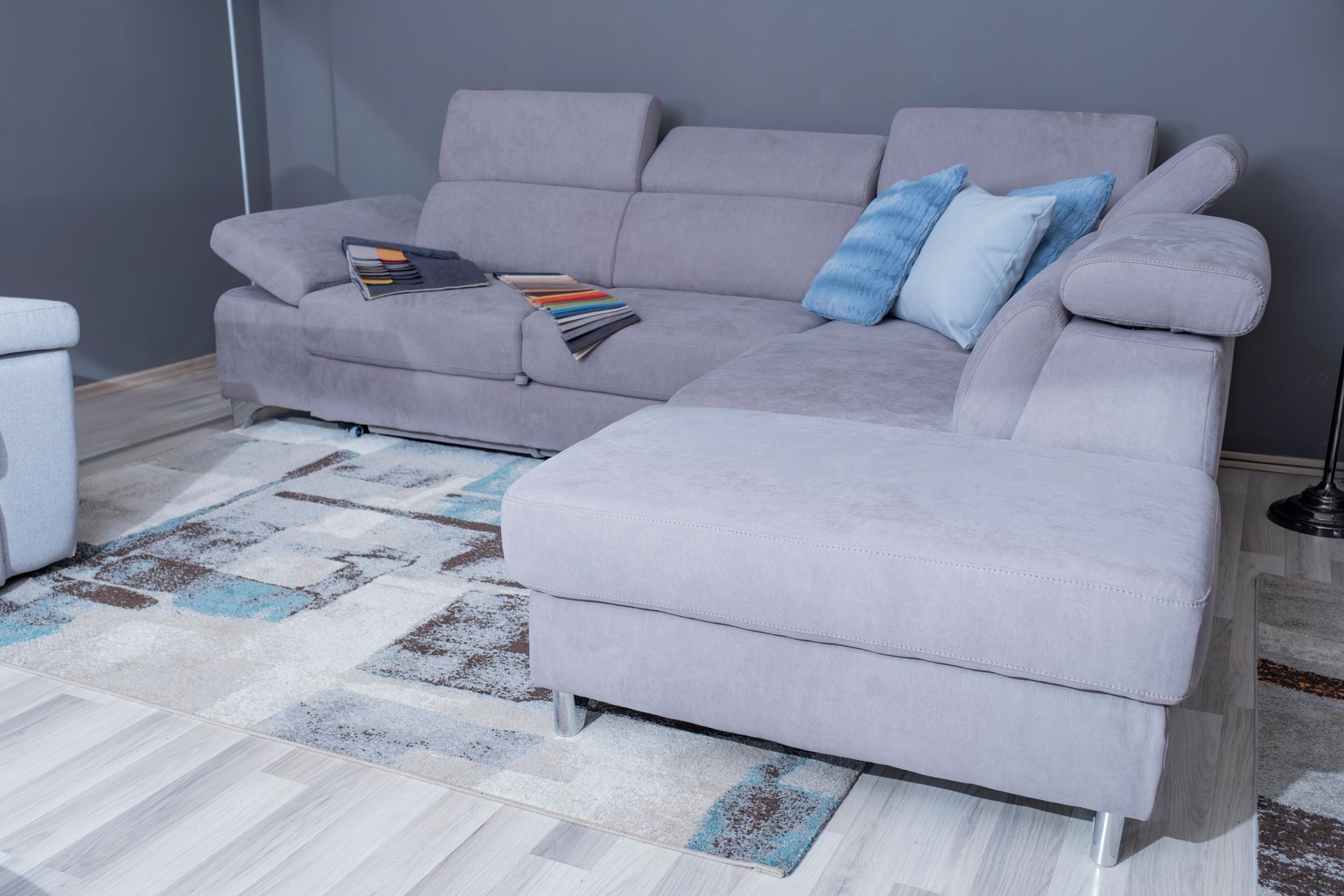 Divano componibile grigio chiaro con chaise longue su un tappeto a fantasia. Due cuscini blu sul divano.