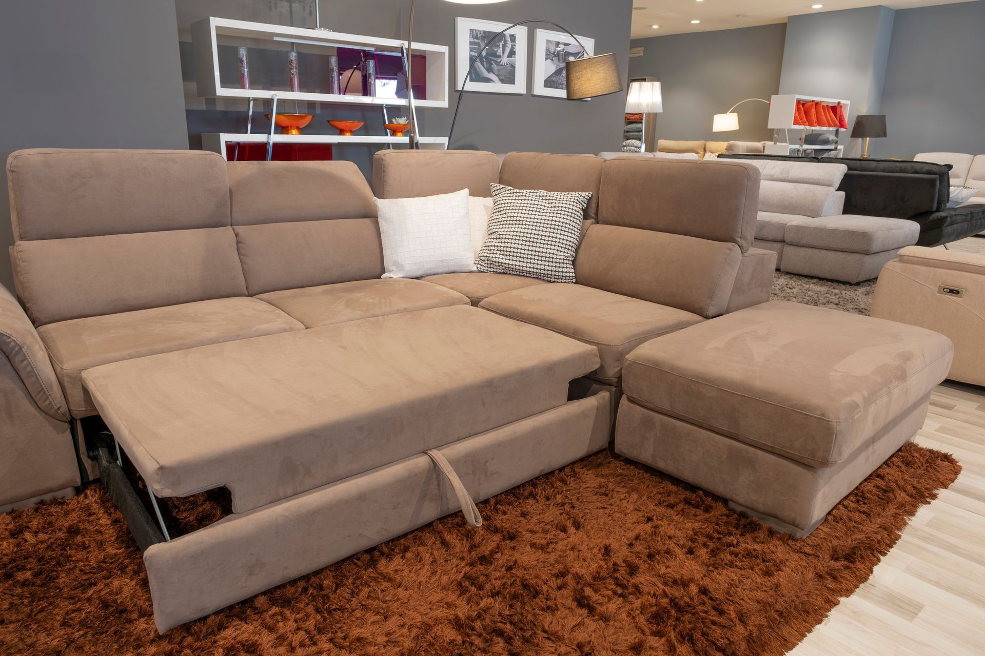 Divano componibile beige con letto estraibile e pouf in uno showroom di mobili, su un tappeto marrone.