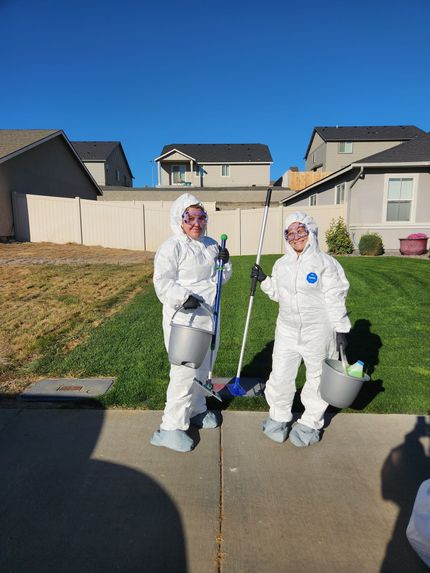 Two people in white protective hazmat suits hold buckets and a long-handled tool while standing on a suburban sidewalk.