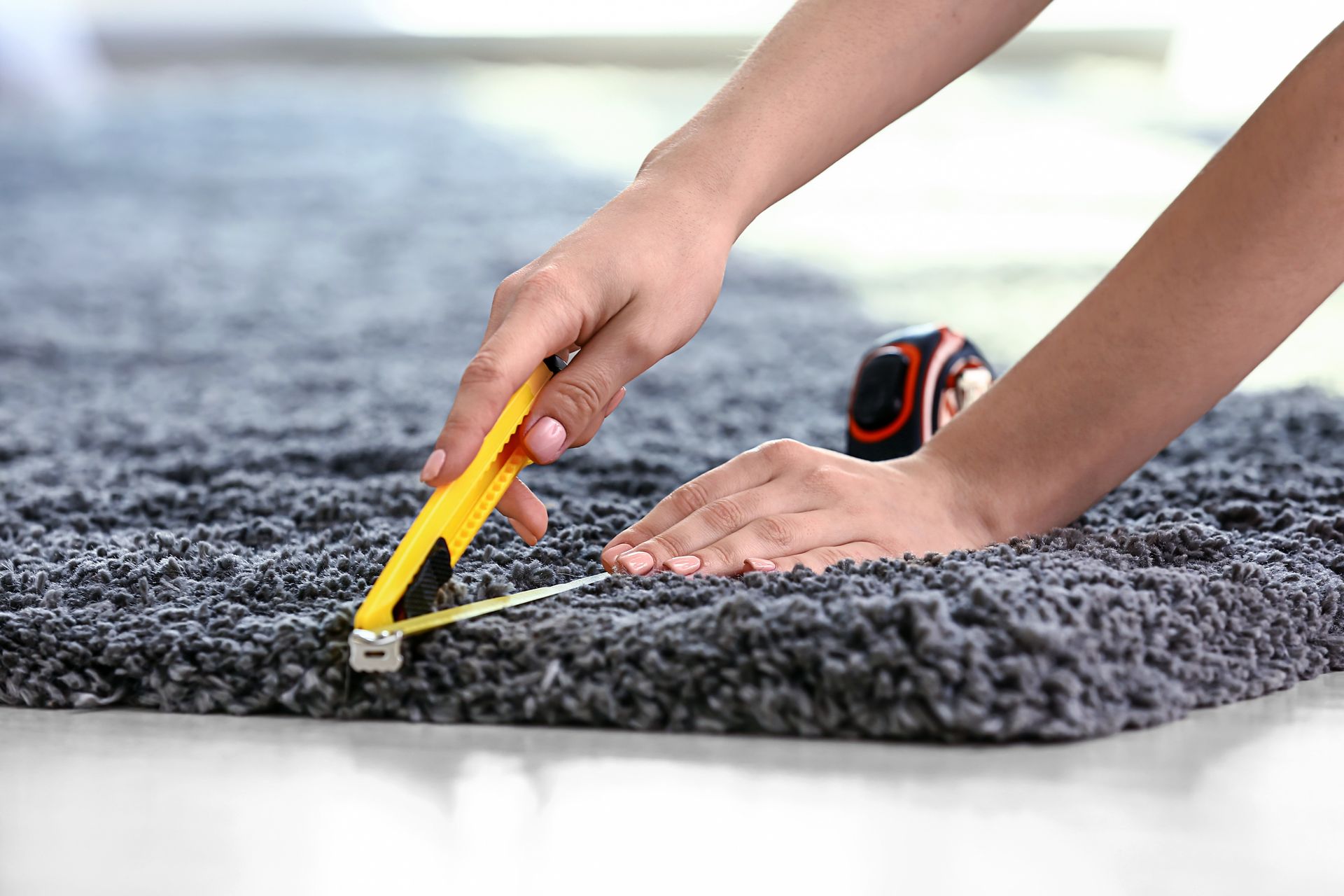 A person uses a utility knife to trim the edge of a thick, dark grey shag rug on a light-colored floor.