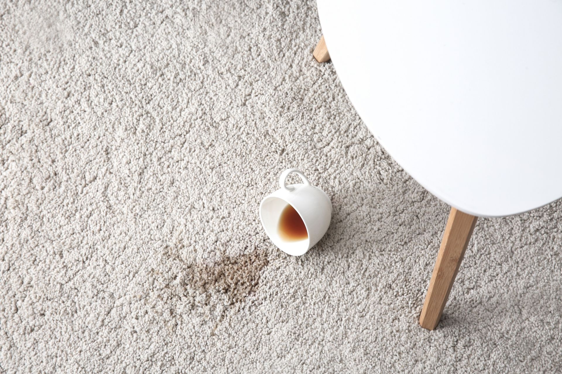 A spilled cup of coffee creates a brown stain on a light-colored, textured carpet next to a white table leg.