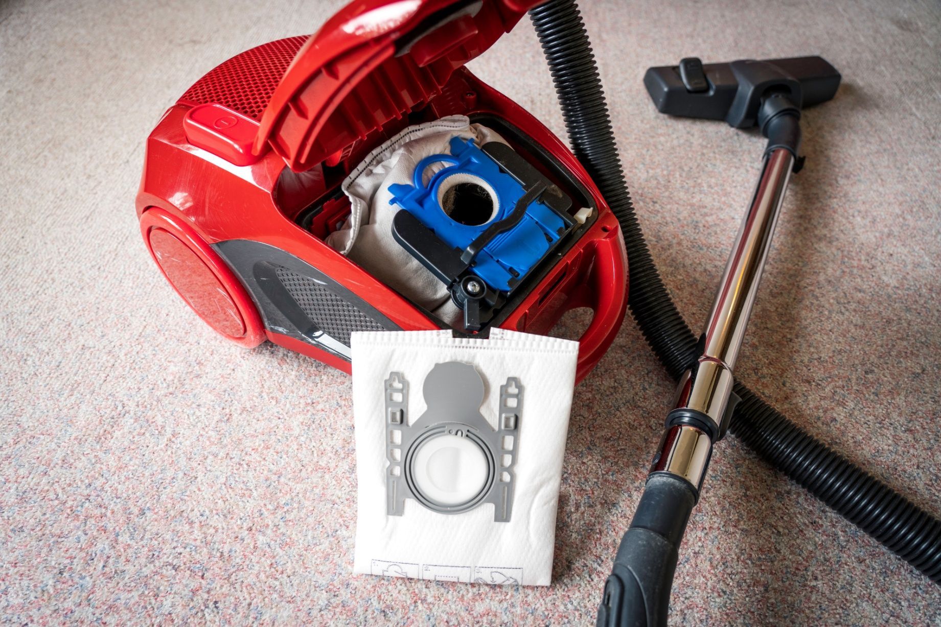 A bright red vacuum cleaner with its lid open, a blue bag holder inside, and a new replacement bag held in the foreground.