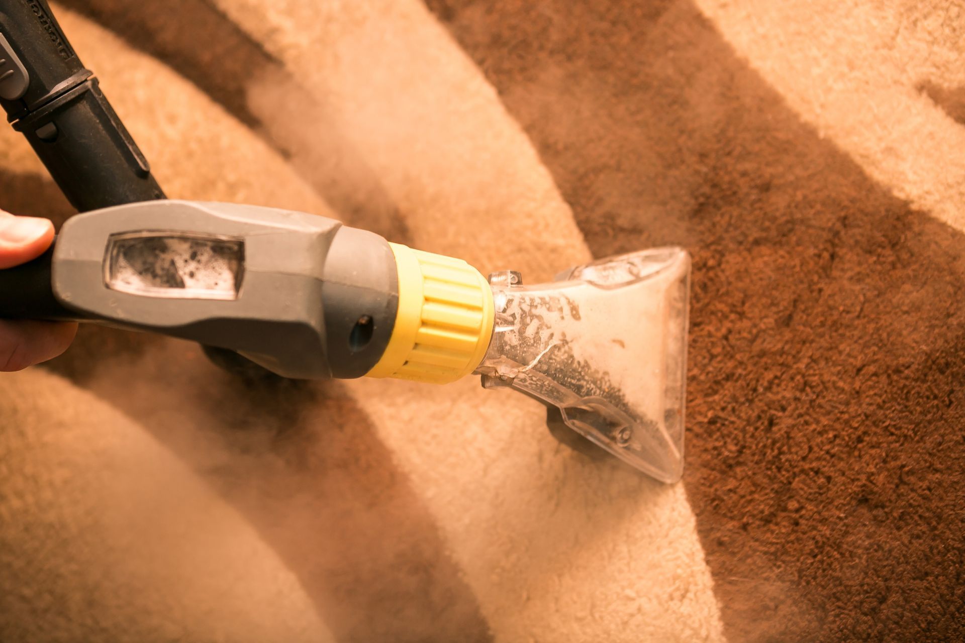 A handheld upholstery cleaner cleans a striped, tan and dark brown carpet, showing a clear contrast between dirty and clean.