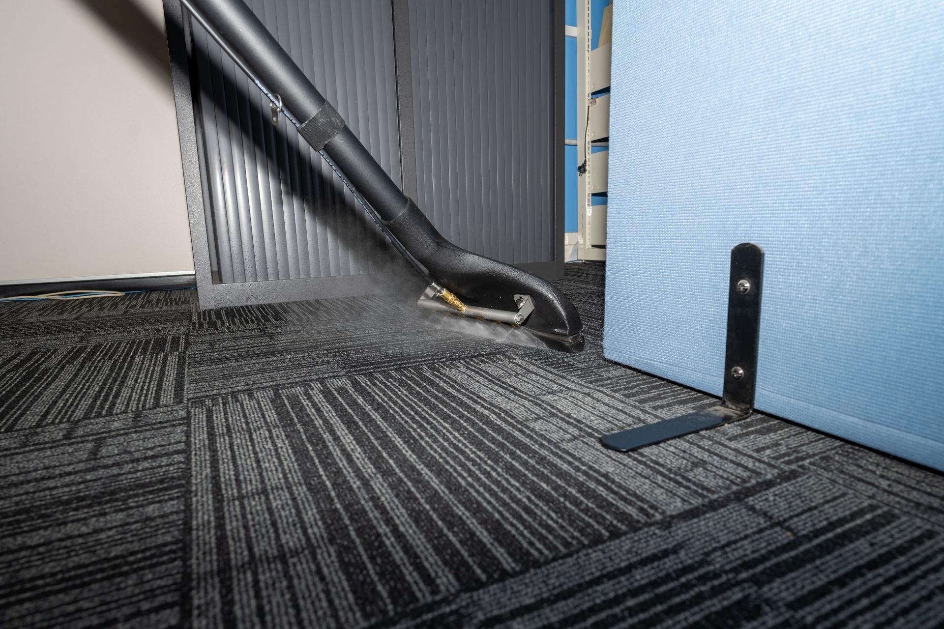 A vacuum cleaner nozzle cleaning a dark, patterned carpet near a blue cubicle wall in an office.