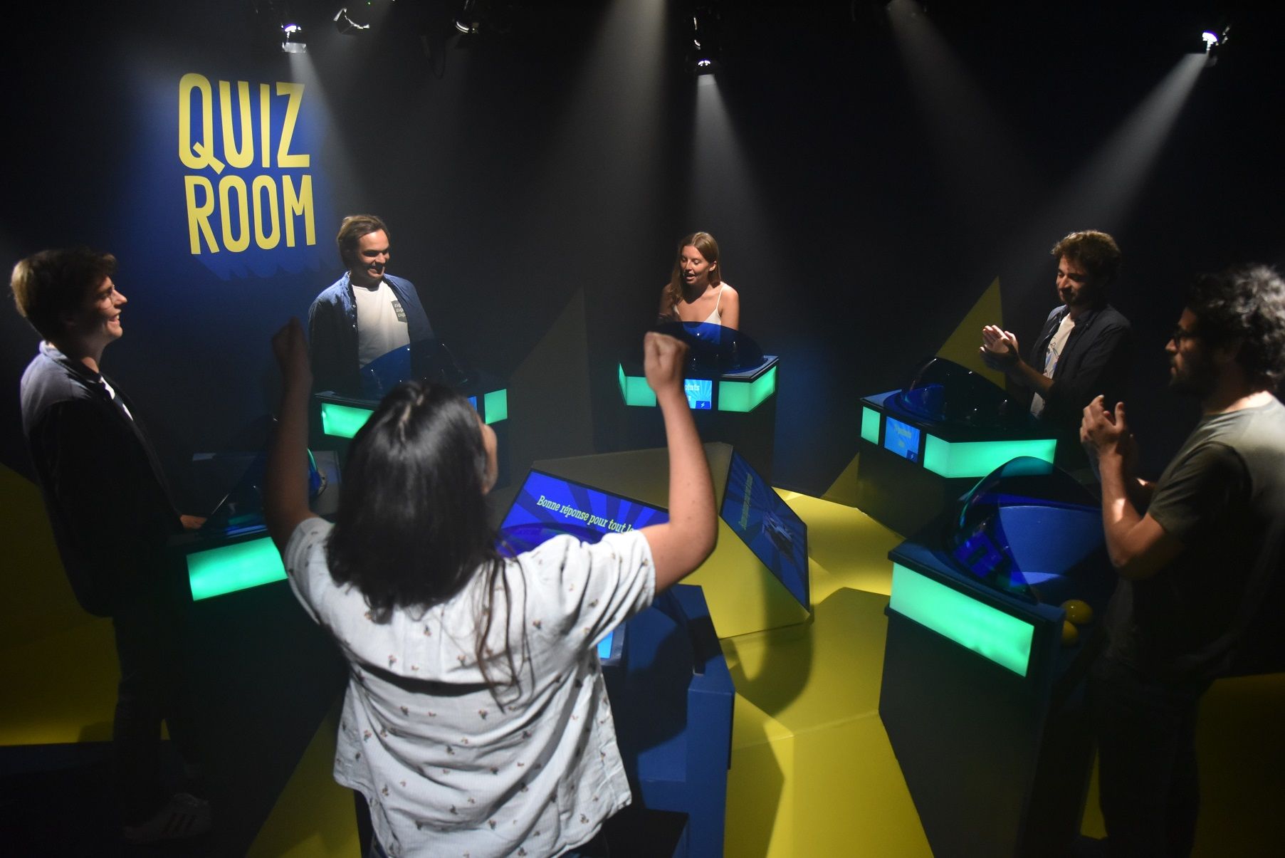 Group of people cheering around a quiz buzzer in front of a blue wall with “QUIZ ROOM” written on it.