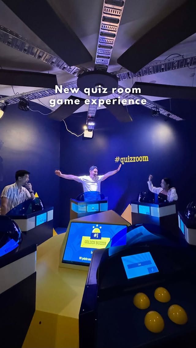 Group of people cheering around a quiz buzzer in front of a blue wall with “QUIZ ROOM” written on it.