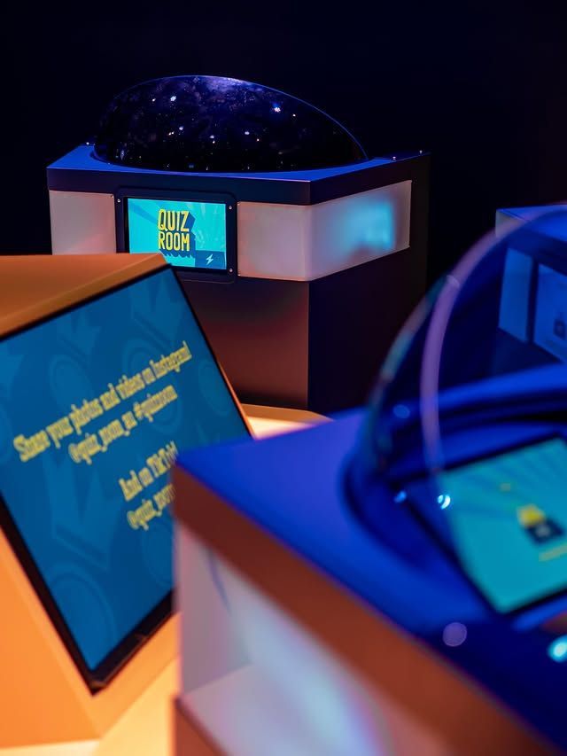 People in a quiz show room. Contestants at podiums, blue and yellow accents, 