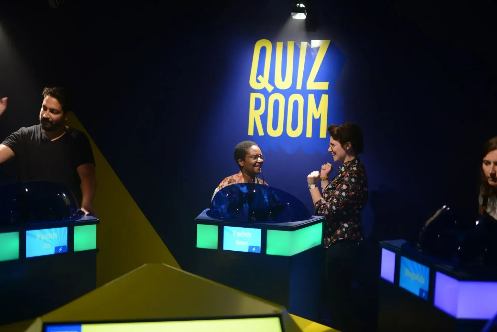 People in a quiz show room. Contestants at podiums, blue and yellow accents, 