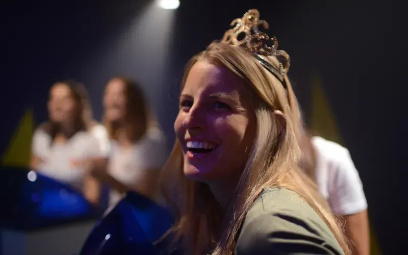 Woman with tiara laughs, illuminated by spotlight. Others in white shirts are blurred in the background.