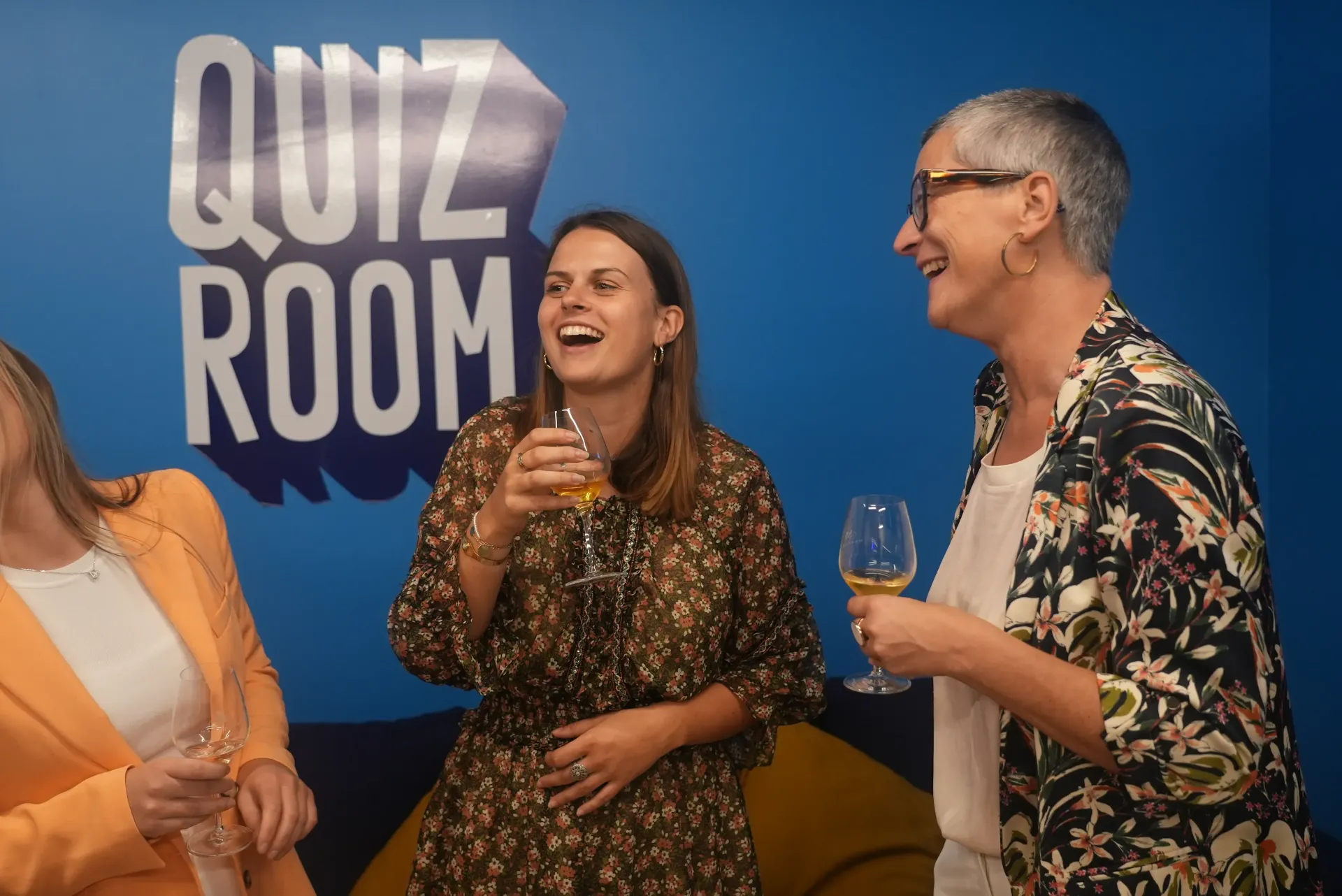 Three women laughing, holding drinks, in front of a "Quiz Room" sign.