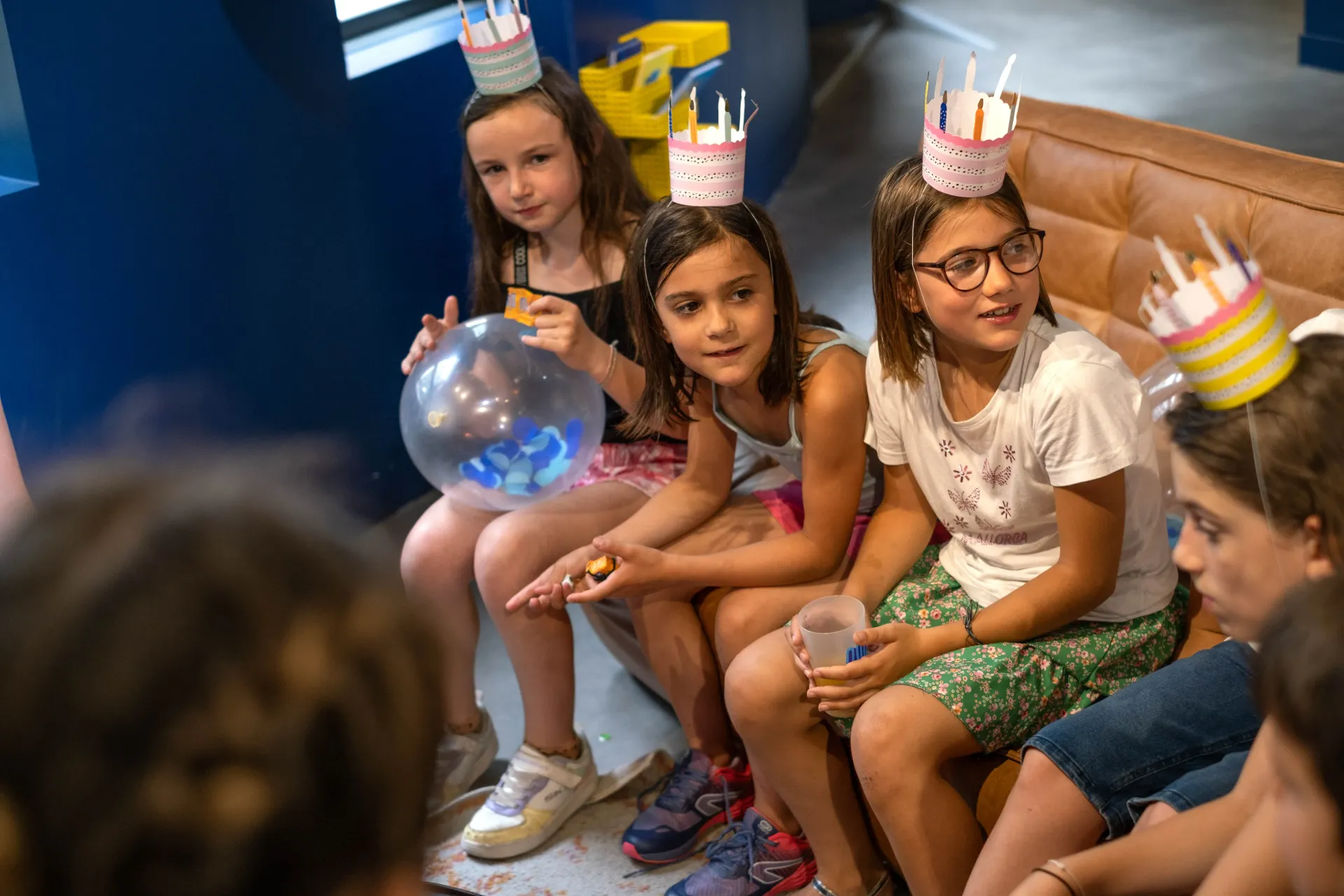 Children with paper crowns sit on a sofa. One holds a glowing balloon. Blue wall in the background.