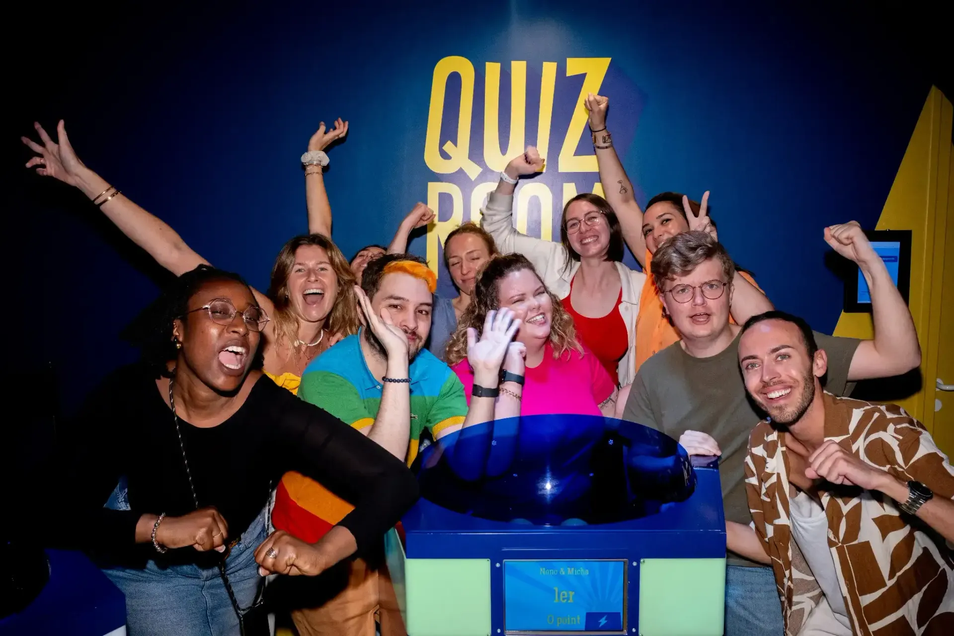 Group of people cheering around a quiz buzzer in front of a blue wall with “QUIZ ROOM” written on it.