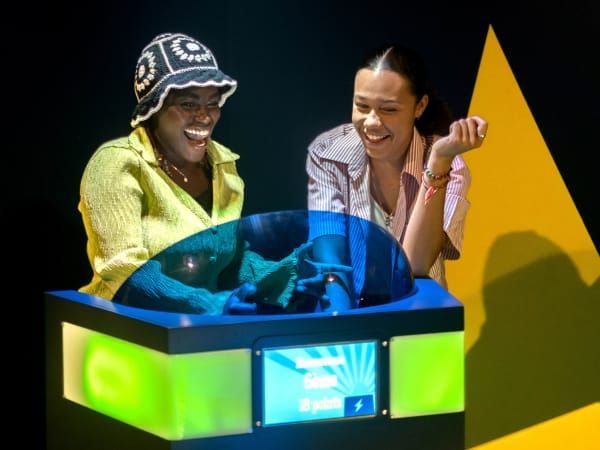 Two people smiling at a brightly lit display. Neon green and blue lights, black backdrop.