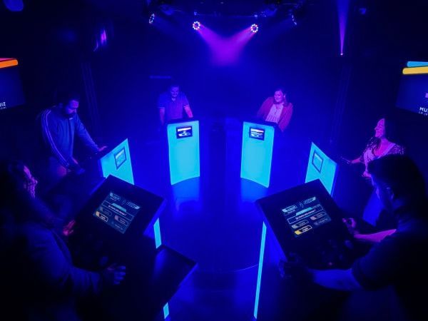 Group of people cheering around a quiz buzzer in front of a blue wall with “QUIZ ROOM” written on it.