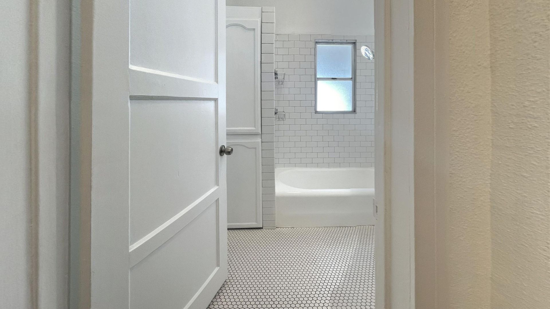 White bathroom doorway opening to a bathroom with a tub, white subway tile, and patterned floor.