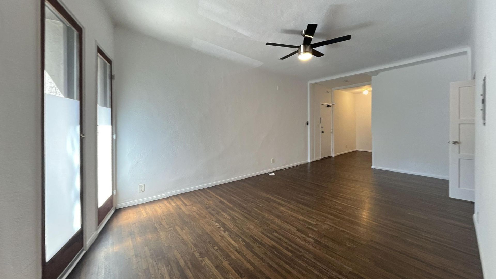 Empty room with hardwood floors, white walls, and a ceiling fan. Two French doors and a hallway.