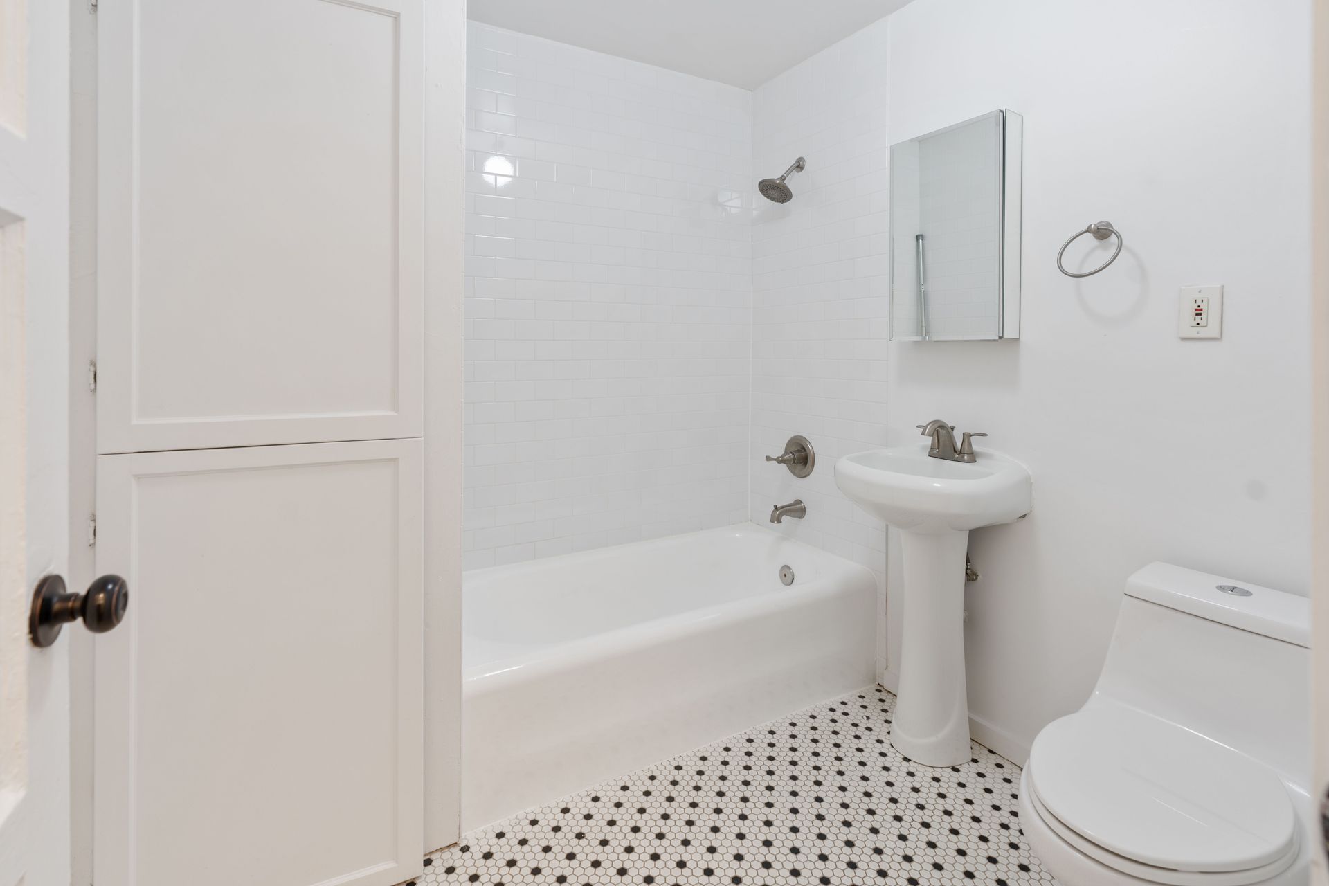 White bathroom with tub, pedestal sink, toilet, and black and white tile flooring.