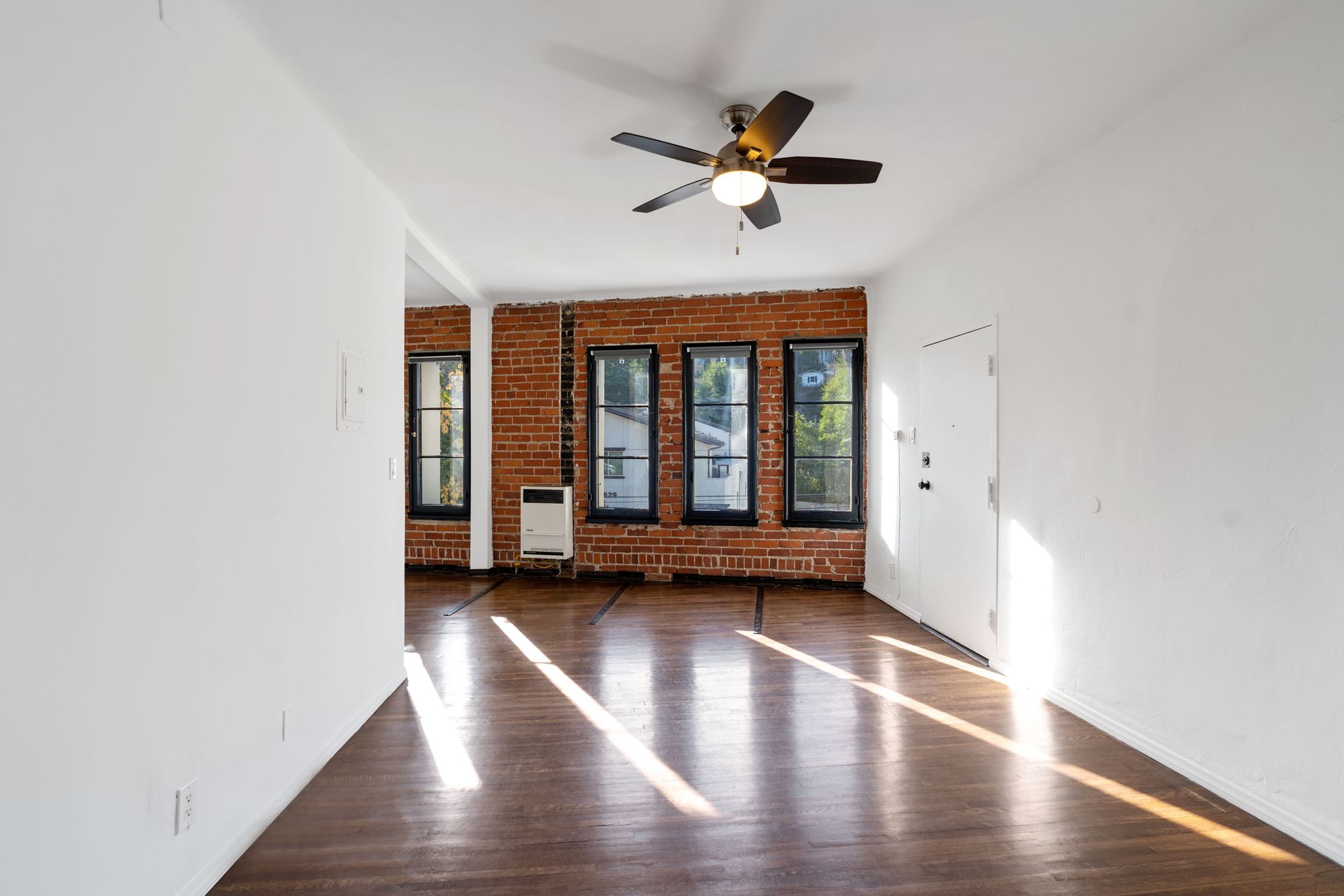 Empty room with hardwood floors, brick wall, three windows, ceiling fan, and white walls.