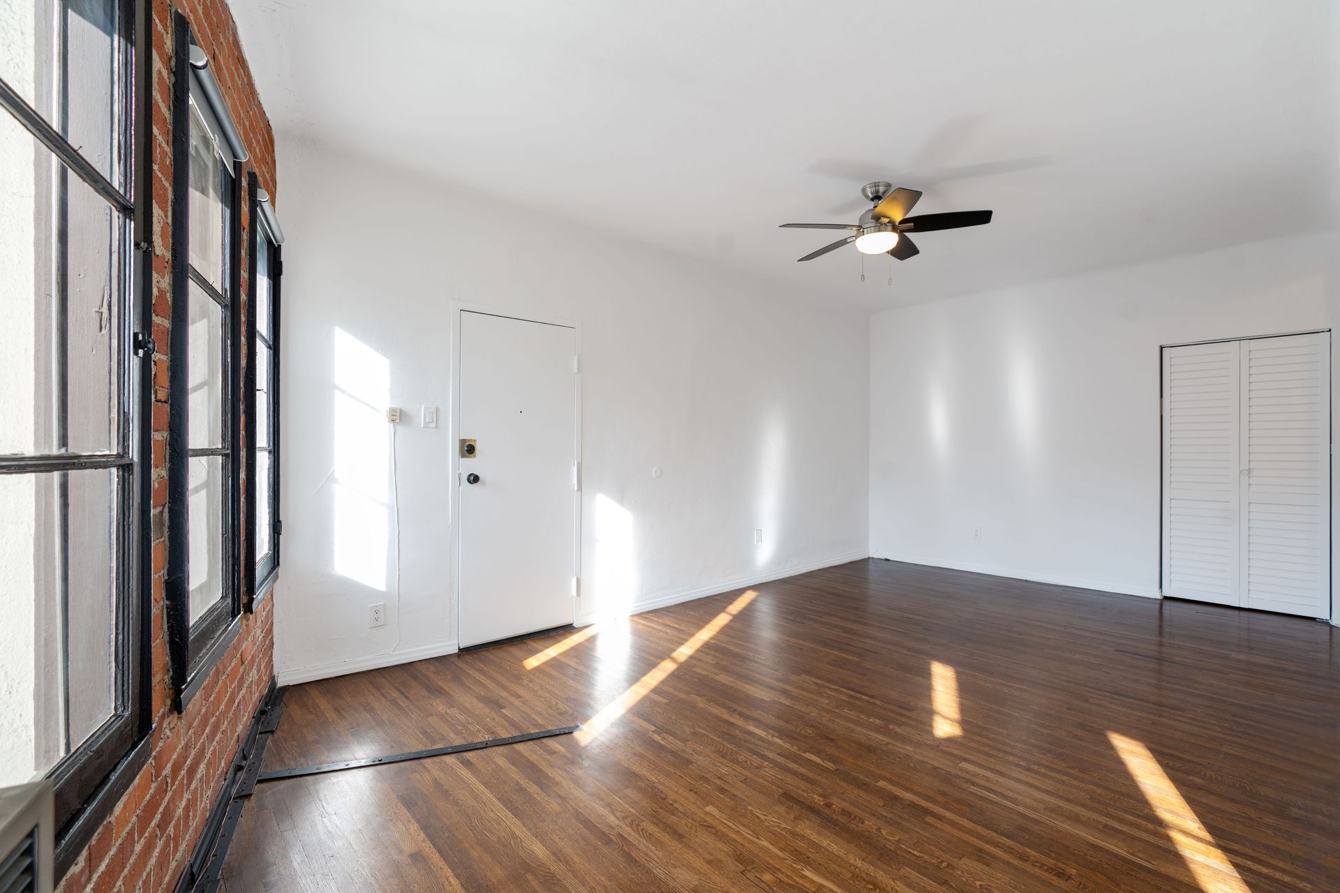 Empty room with hardwood floors, white walls, large window, brick wall, and ceiling fan.