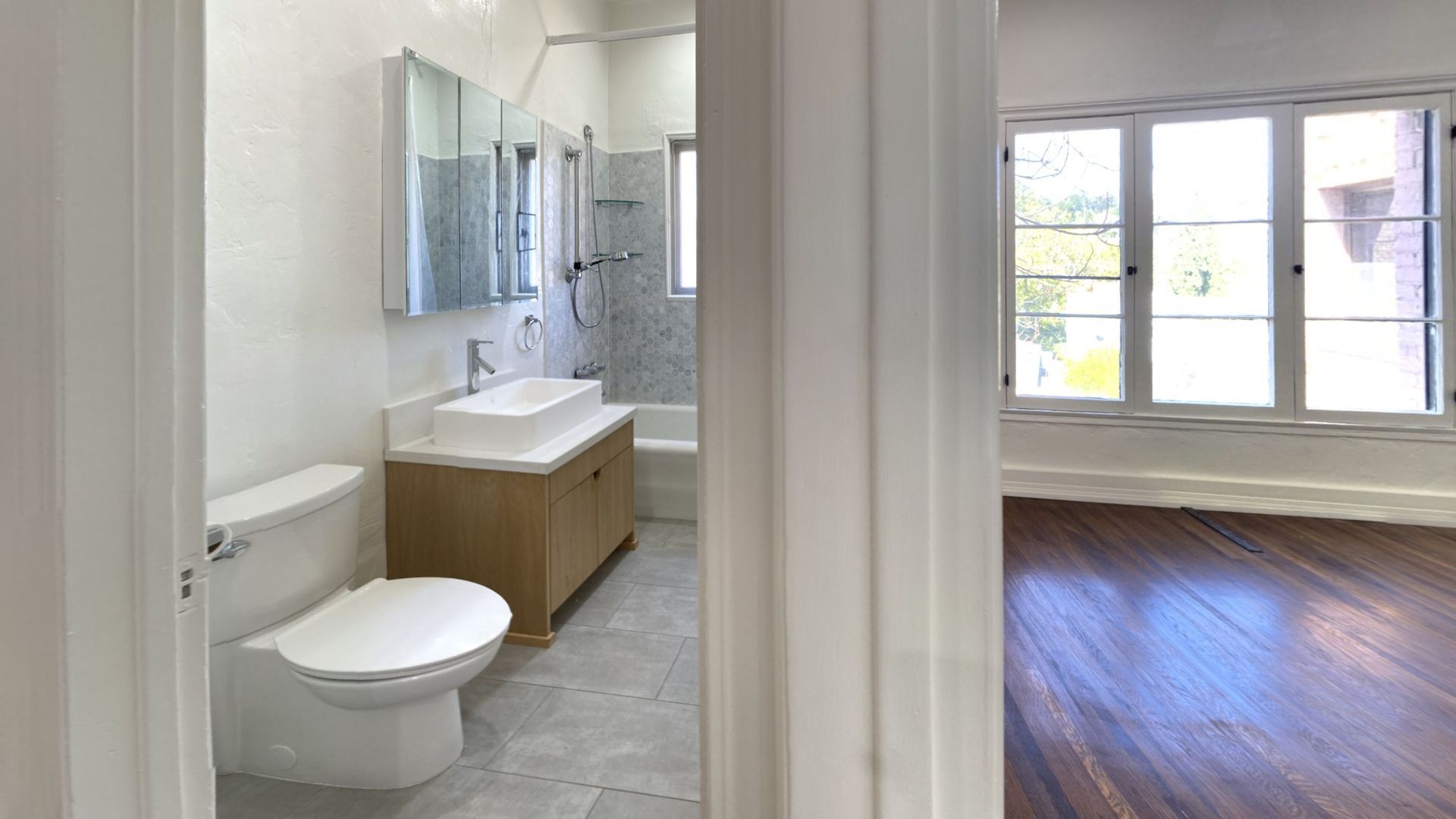 Bathroom with white walls, toilet, vanity, and a glimpse into a room with hardwood floors and large windows.