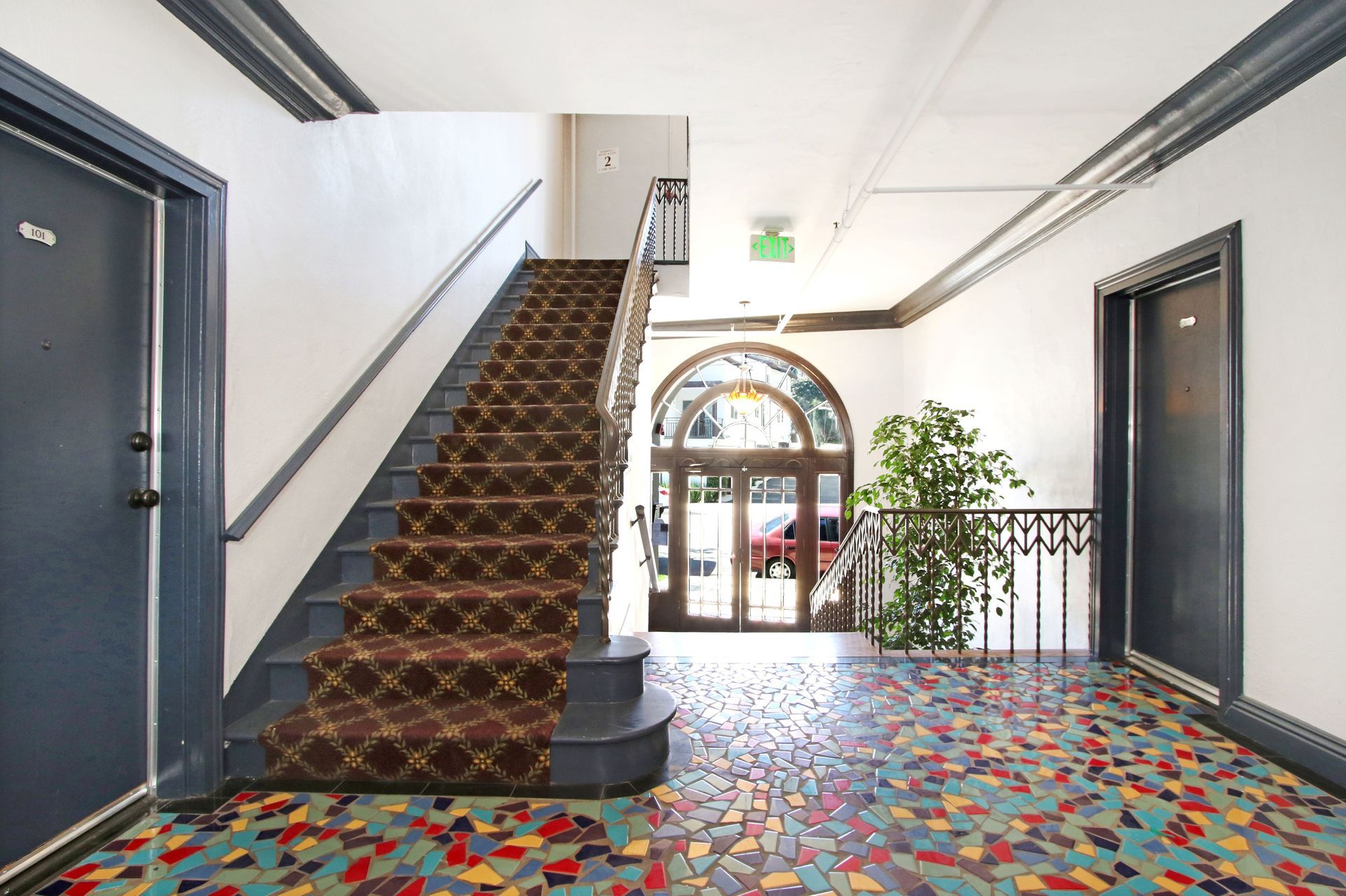 Apartment building hallway with staircase, mosaic tile floor, and arched doorway to outside.