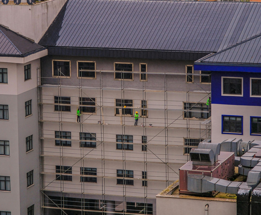 Construction workers in bright vests work on scaffolding outside a multistory building under a dark roof.