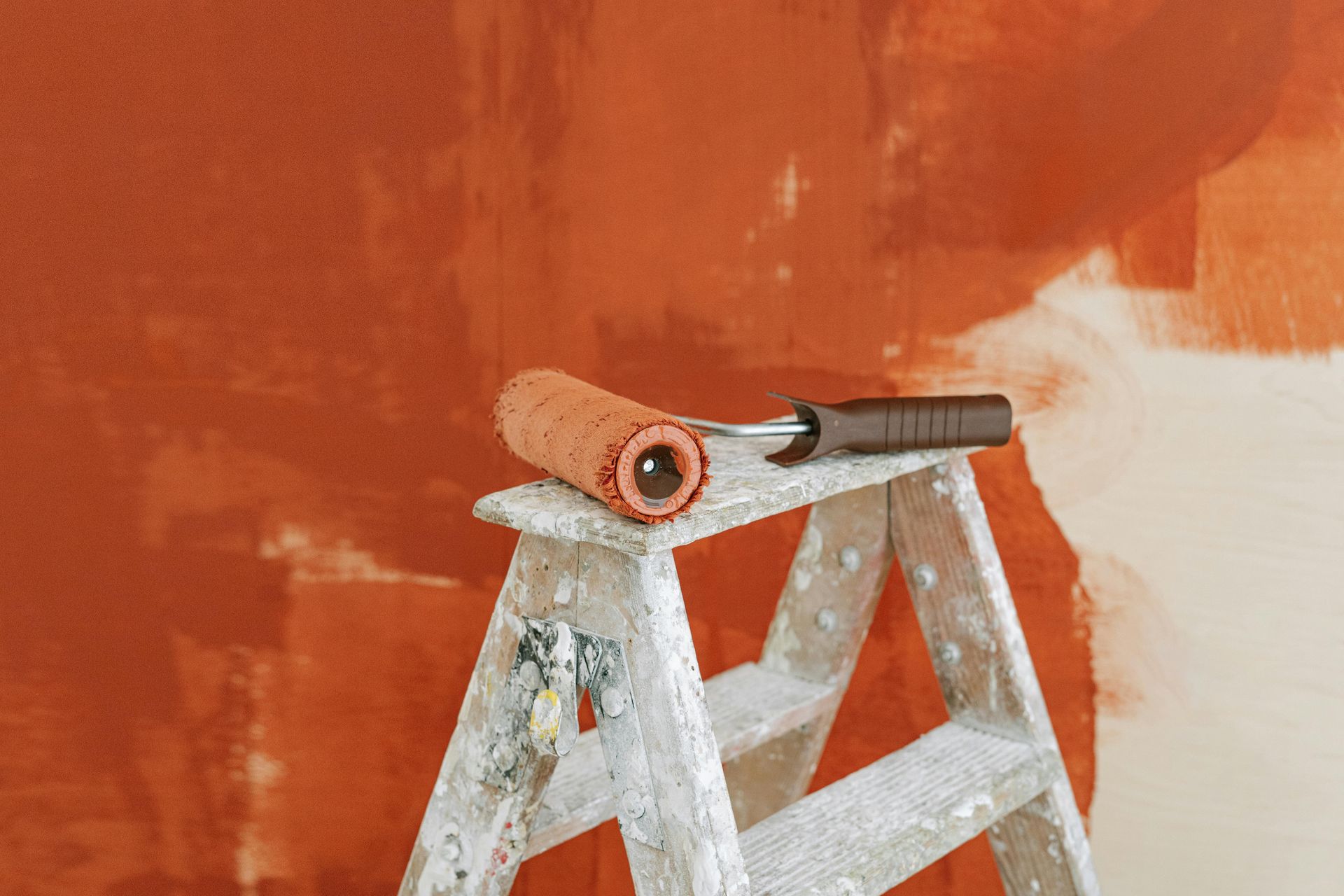 A paint roller coated in orange paint rests on top of a step ladder against a partially painted orange wall.