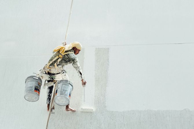 A rope-access worker in safety gear hangs from ropes against a white building wall under a bright blue sky.