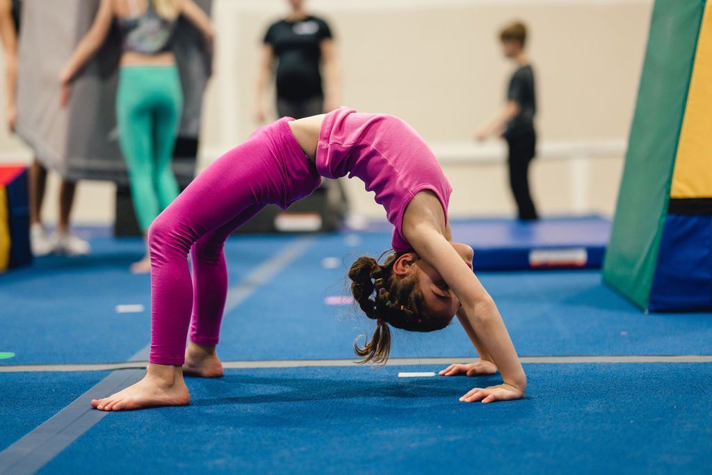 Girl in pink athletic wear doing a bridge on a blue mat in a gymnastics studio.