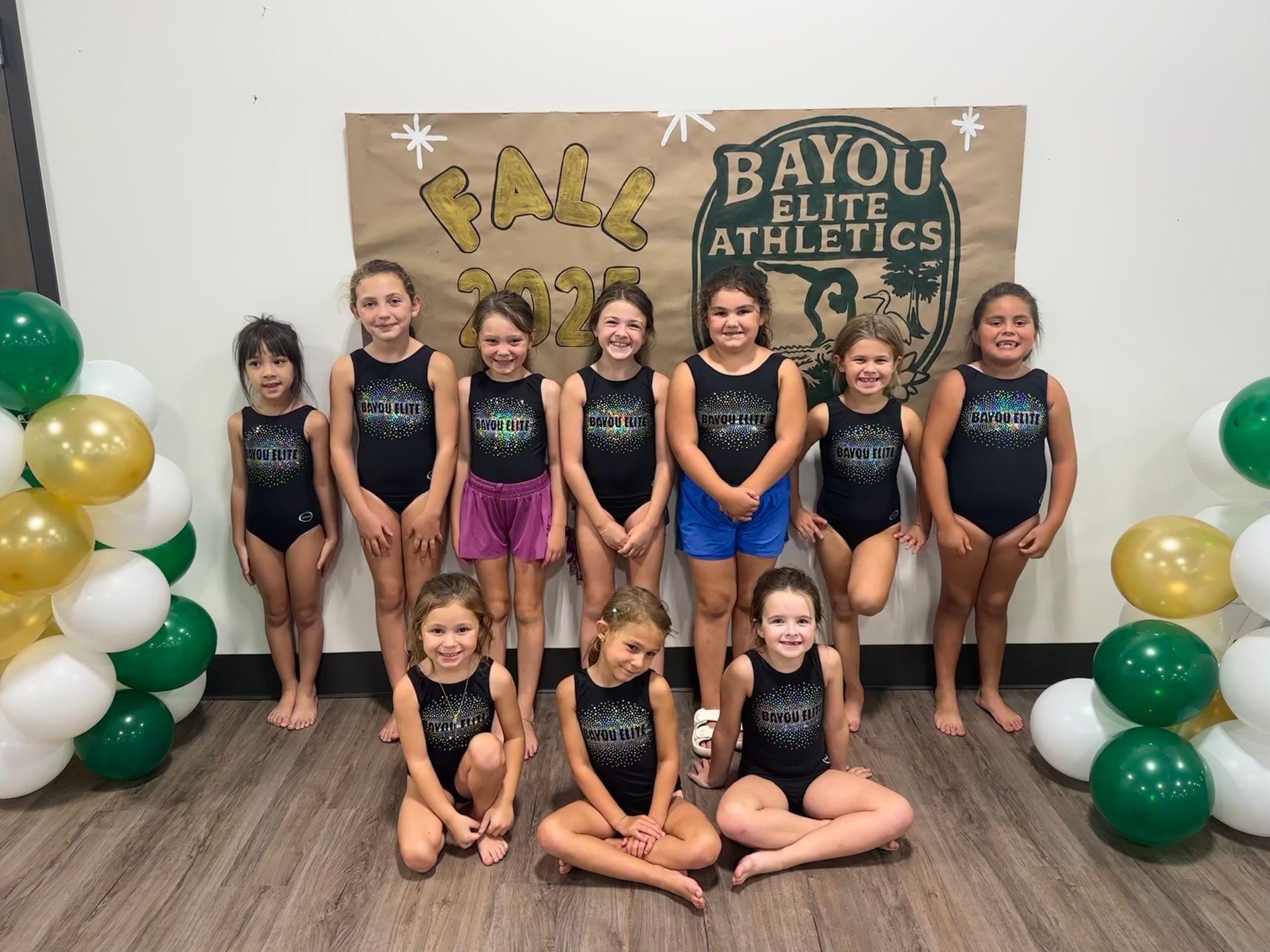 A young Asian girl in a green shirt does the splits on a blue mat, smiling at the camera.