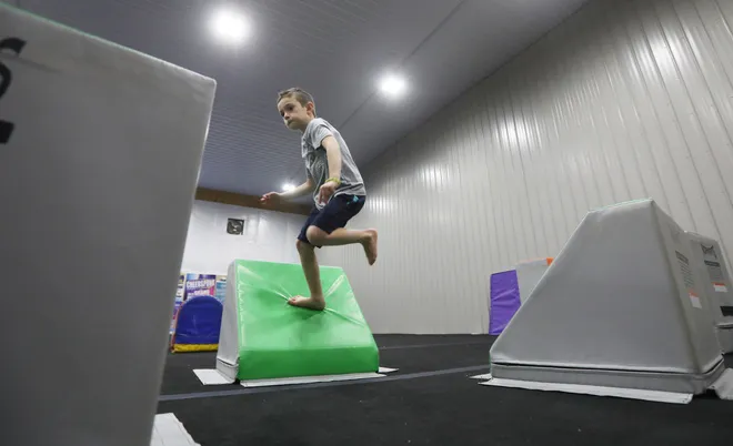 A young boy in a gym is mid-jump over a green, padded cube. He is wearing a t-shirt, shorts, and appears to be barefoot.