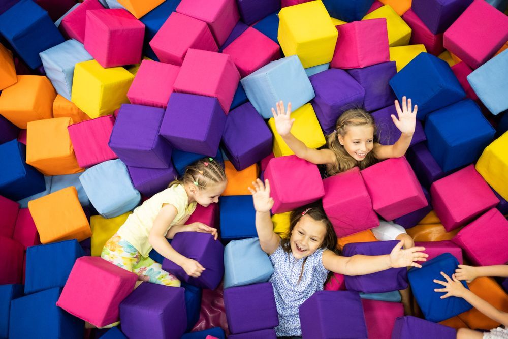 Children playing in a colorful foam cube pit, laughing and raising their arms.