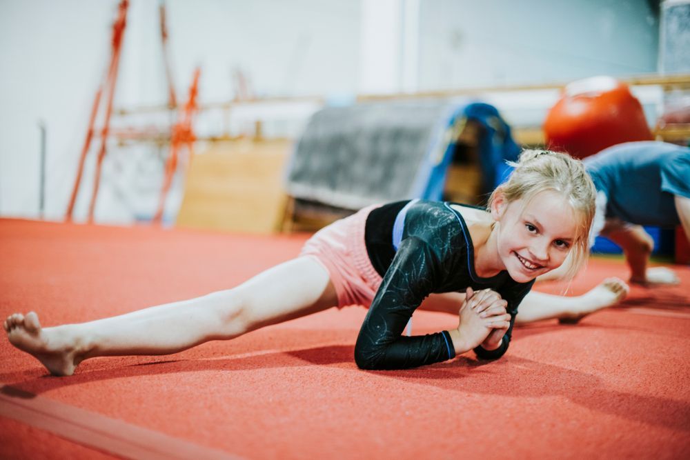 A young girl in a gymnastics leotard smiles, performing a split on a red mat in a gym, with other children in the background.