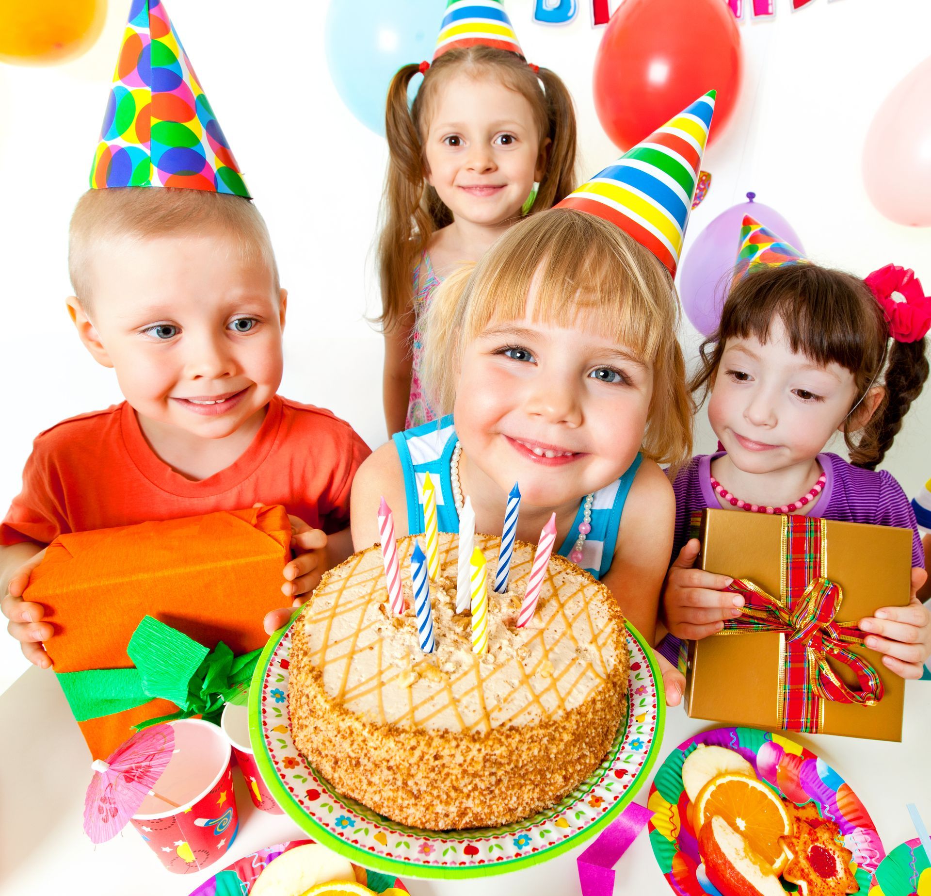 Four children smiling at a birthday cake with candles and gifts in a party setting with balloons and party hats.