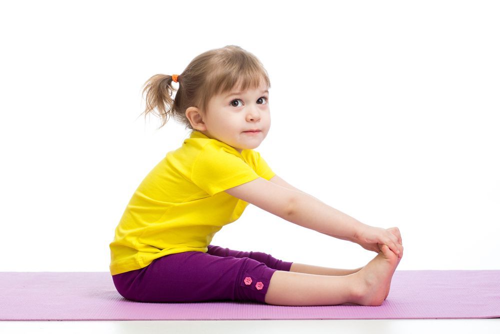 Young child with a yellow shirt and purple pants, stretching on a pink mat, reaching for their toes. White background.