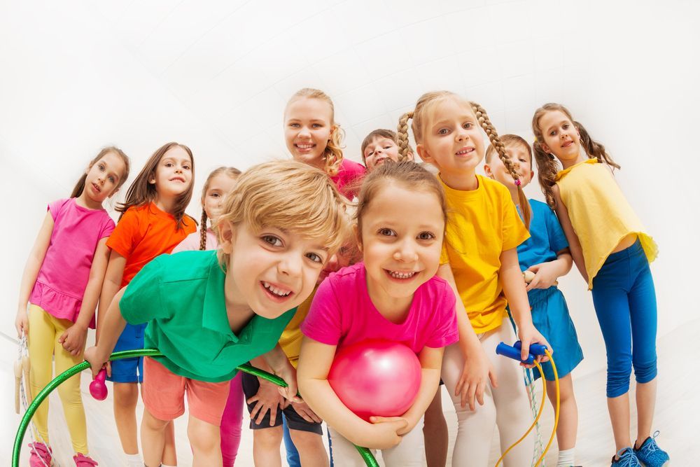 Group of children smiling at the camera. They are holding jump ropes, a ball, and hula hoops, and wearing colorful clothes.