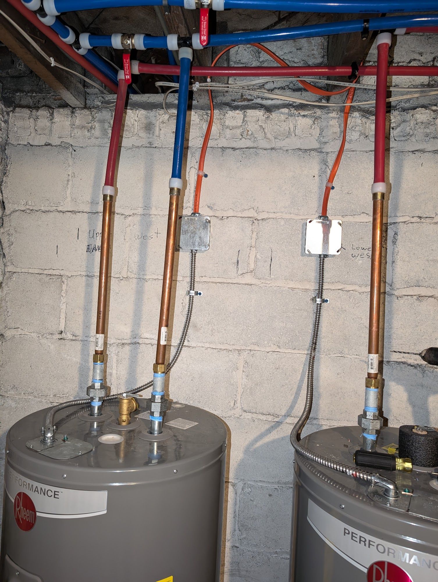 Two water heaters with copper and blue/red pipes against a cinder block wall.